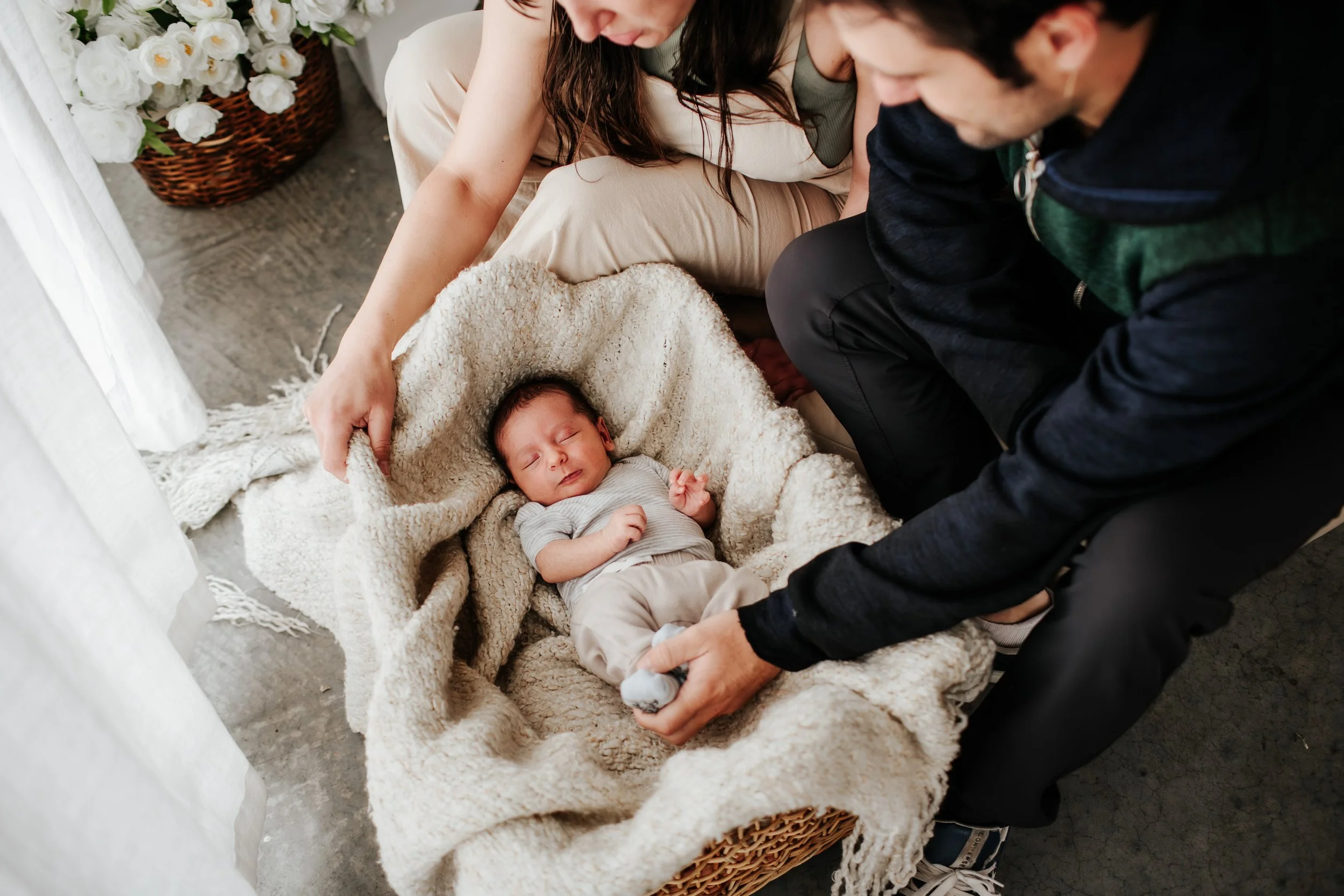 A baby sleeping in a cozy blanket with two adults attending to him, near a basket of white flowers.