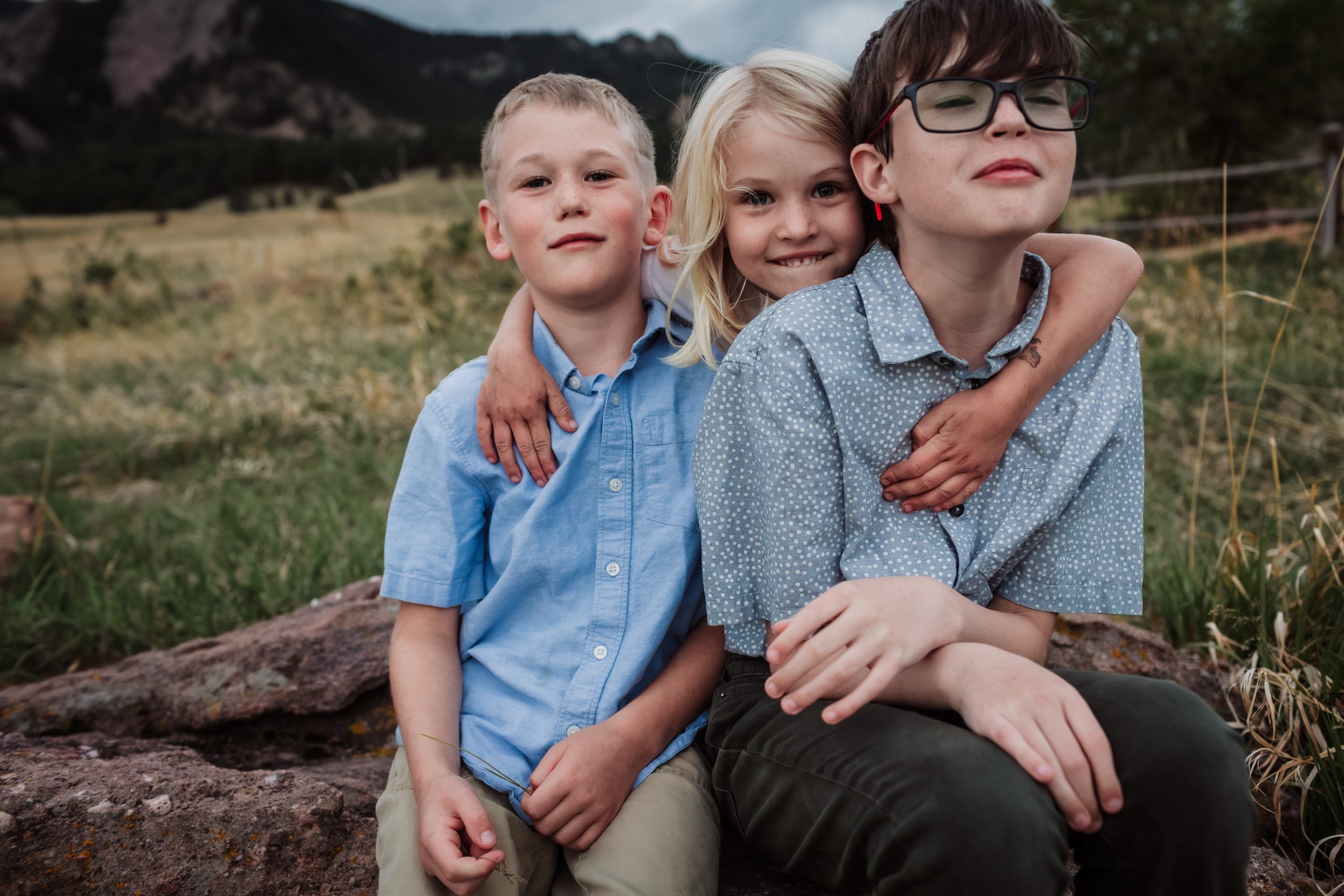 Three children sitting outdoors on a rock with a mountain landscape in the background. They are smiling and hugging each other, with one boy wearing glasses on the right, a girl in the middle, and a boy on the left.