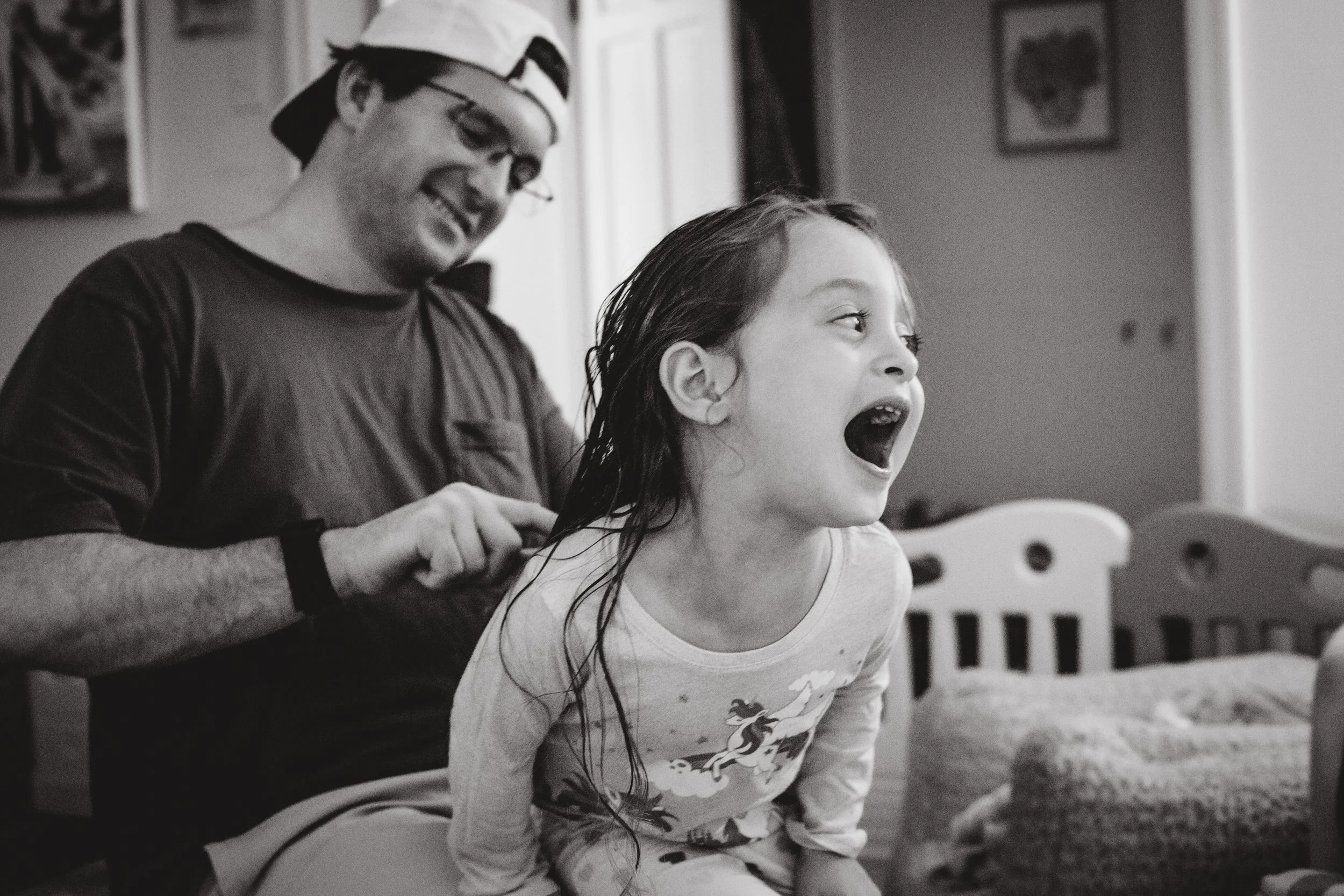A man and a young girl sitting together, smiling and laughing. The man is wearing glasses and a cap backwards, while the girl has long hair and is wearing a T-shirt with a unicorn design. They are in a cozy room with framed pictures on the wall.