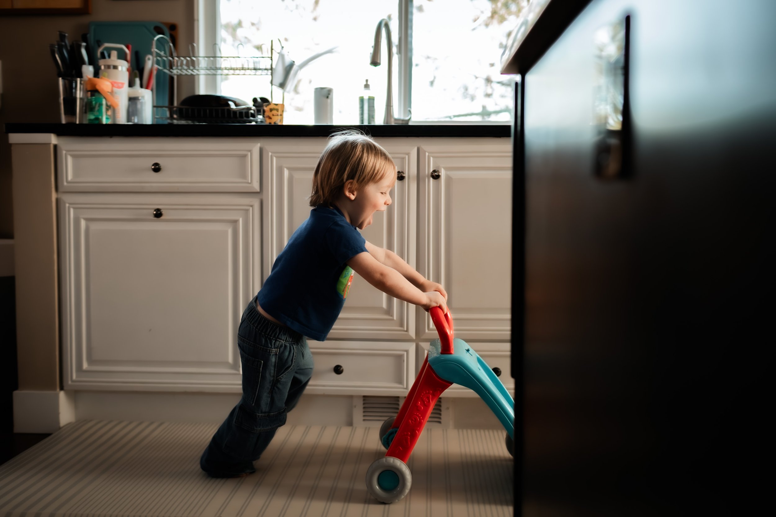 A young boy with blonde hair playing with a toy stroller in a kitchen with white cabinets and a window in the background.