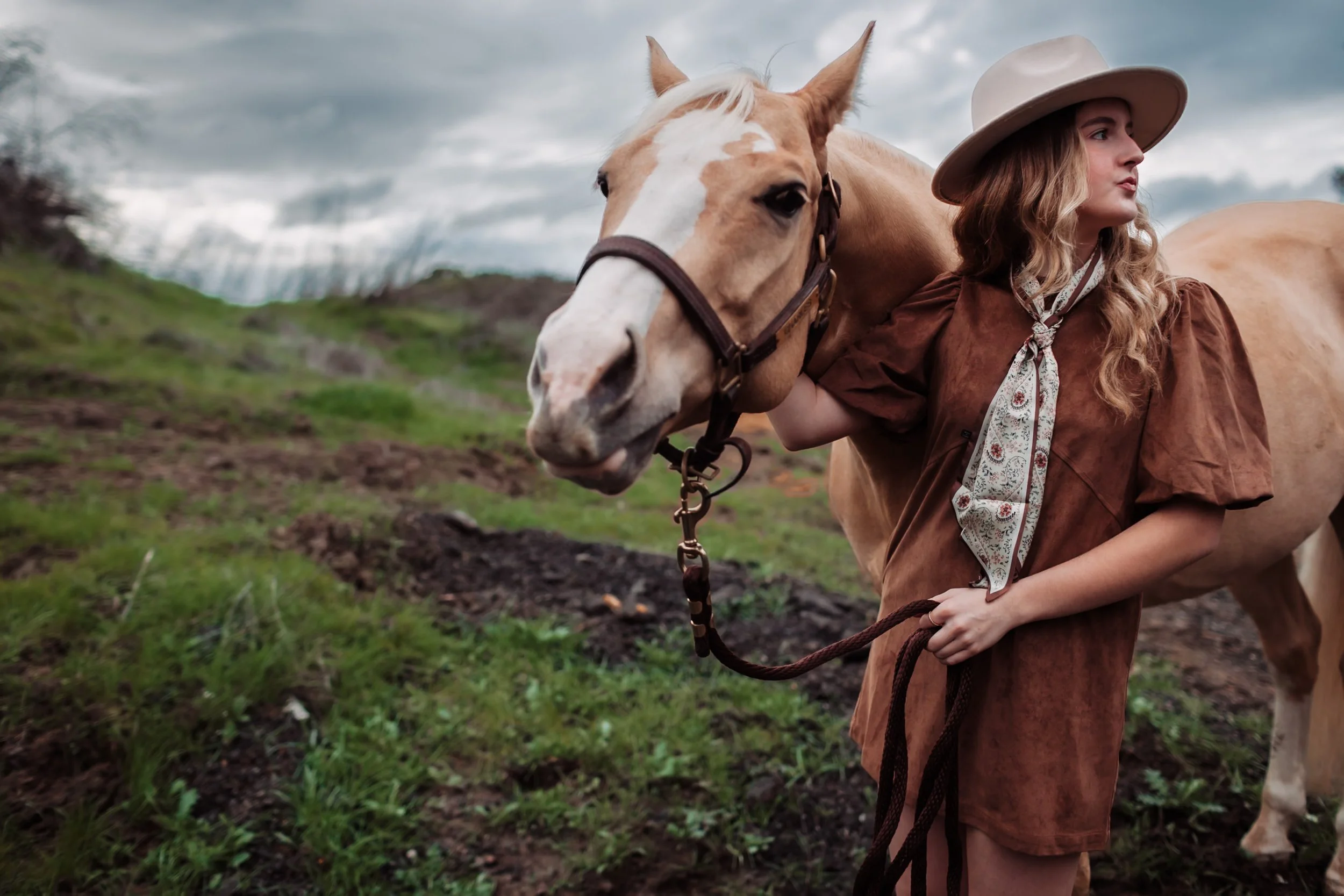 A woman with long curly hair wearing a wide-brimmed hat and brown outfit stands beside a cream-colored horse with a white mane in a grassy outdoor area under a cloudy sky.