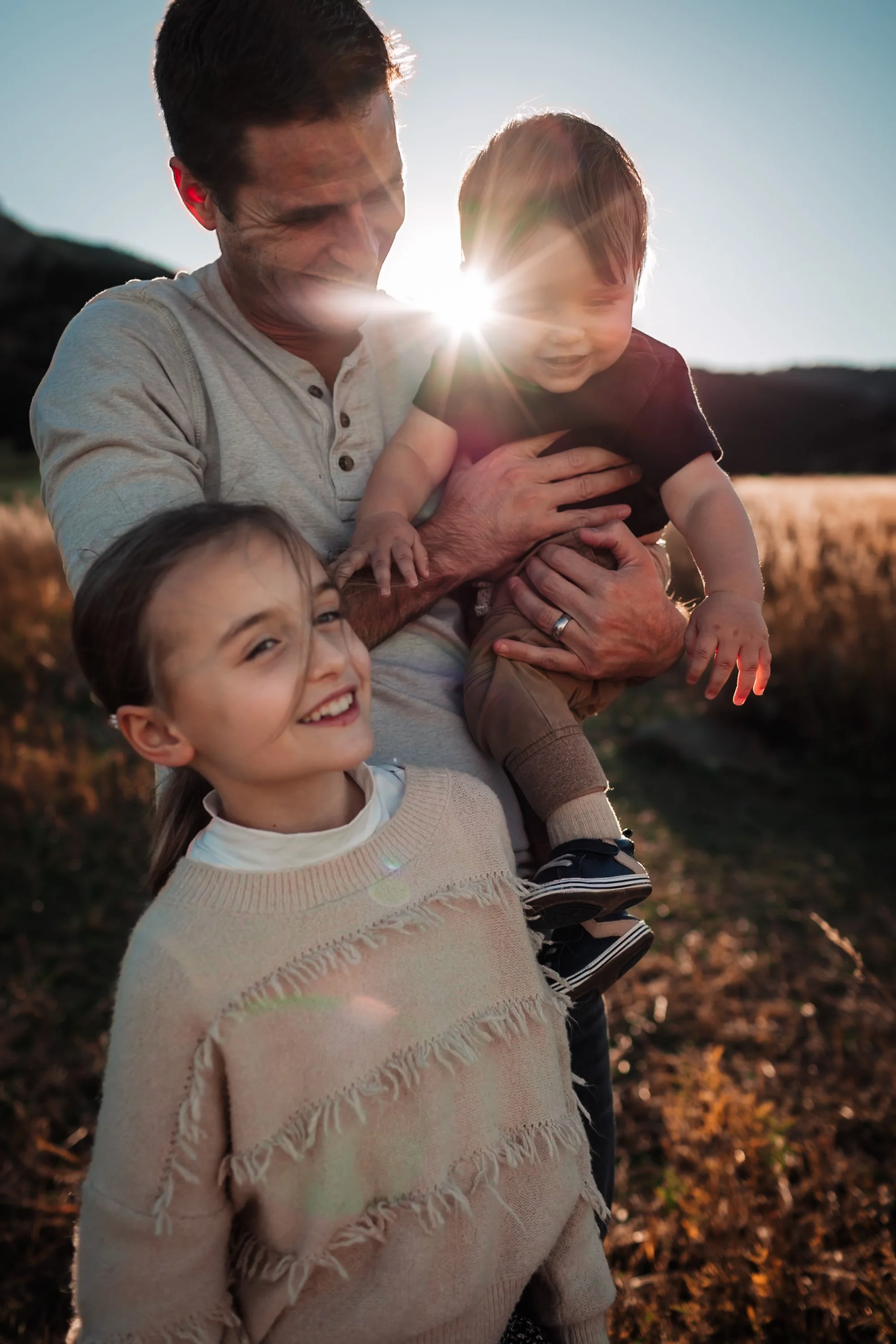 A man holding a young boy on his shoulder, both smiling and laughing outdoors during sunset, with warm sunlight creating a lens flare and backlit effect.
