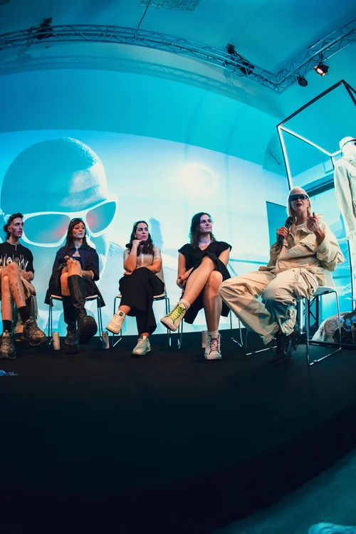 A group of people sitting on chairs in front of a large screen displaying an image of a person's face wearing white sunglasses and a hat, with a bright background. The setting is a panel for fashion brand Oakley featuring Jocelyn Burnham