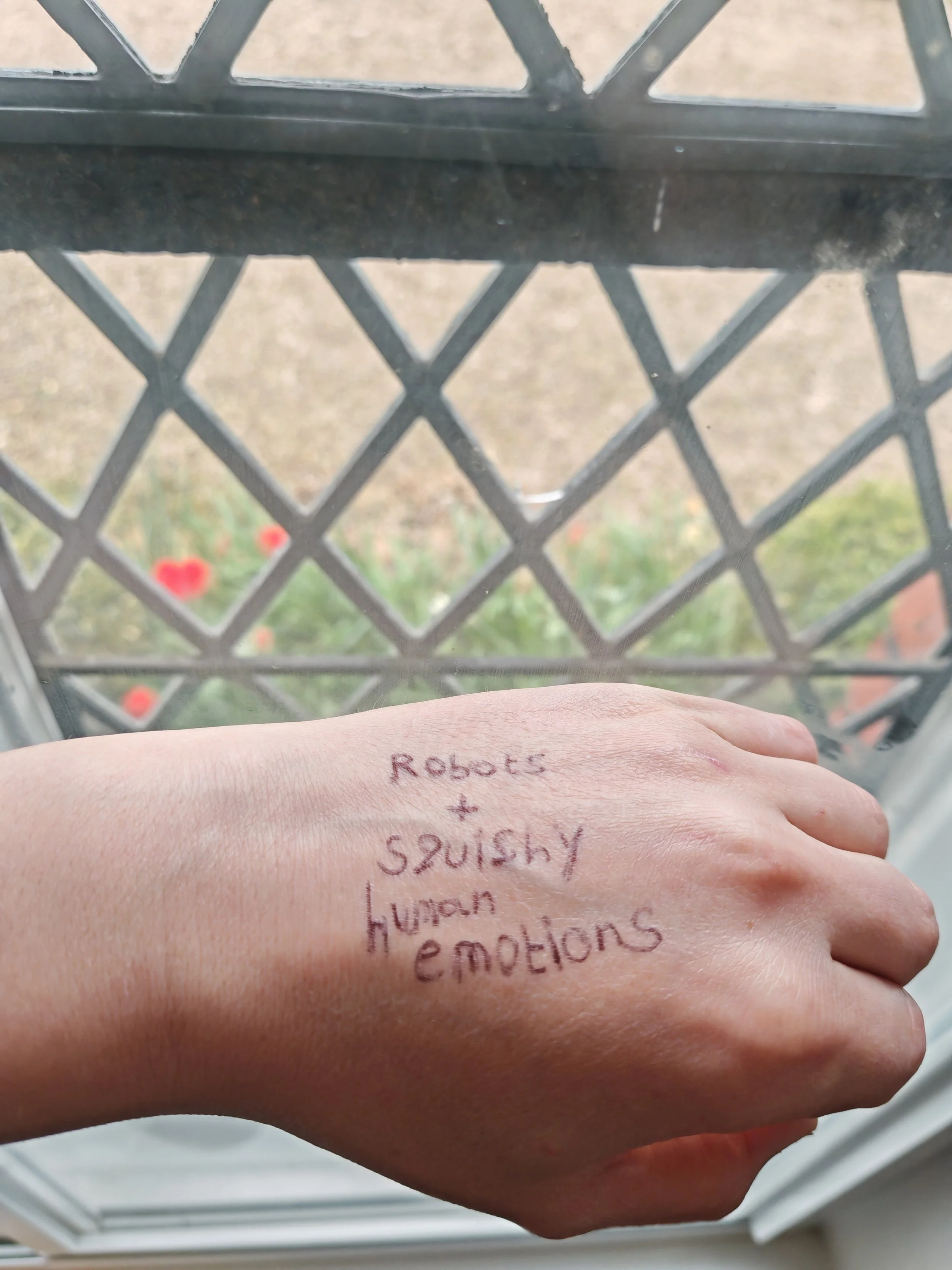 A photo of the back of Jocelyn's hand, with the words "Robots + squishy human emotions" written on it with a biro pen
