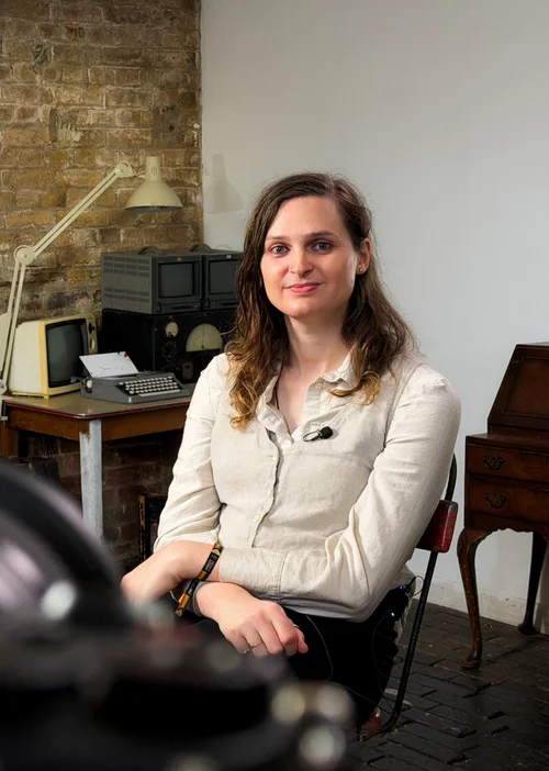 A woman (Jocelyn Burnham) with long brown hair wearing a light-colored shirt sitting on a chair in a room with a brick wall and vintage electronics, including a small TV, radio, and typewriter, in the background.