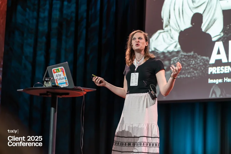 A high energy photograph of Jocelyn Burnham presenting at a conference for Tixly in Oslo, Norway. She is wearing a white dress, a black shirt and has long hair. Her arms are out to the side in an expressive and emotive pose.