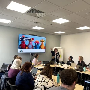A group of people seated around a conference table, listening to a presenter beside a screen showing a slide about co-design