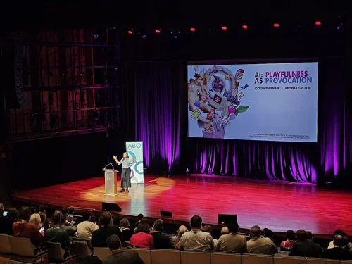 Jocelyn Burnham on a large stage presenting to a seated audience, with a slide titled AI as Playfulness and Provocation on the screen behind her.