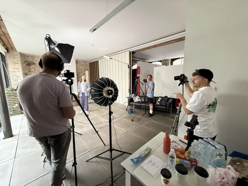 Behind the scenes at a Bookboon filming shoot: crew operating cameras and lighting in a brick-walled room with large windows while Jocelyn Burnham speaks to camera.