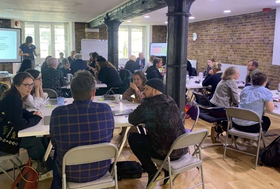 A group of people seated at long tables in a conference room with brick walls and large windows, attending a workshop or seminar.