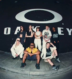 Six young designers standing and sitting against a large black wall with a white logo, outdoors on a city pavement