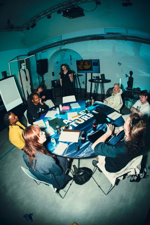 A group of diverse people sitting around a circular table in a dimly lit room with technology and artwork on the walls, some engaged in conversation and others using laptops or holding drinks.
