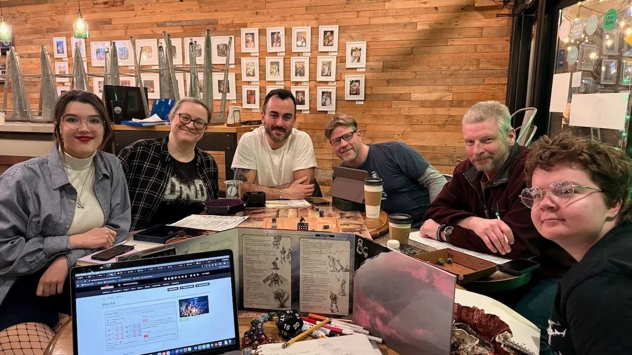 Six people gathered around a table in a restaurant or cafe, playing a tabletop role-playing game. The table has various game materials, including dice, character sheets, and a laptop. The background features a wooden wall with framed pictures, and the atmosphere appears cozy and casual.