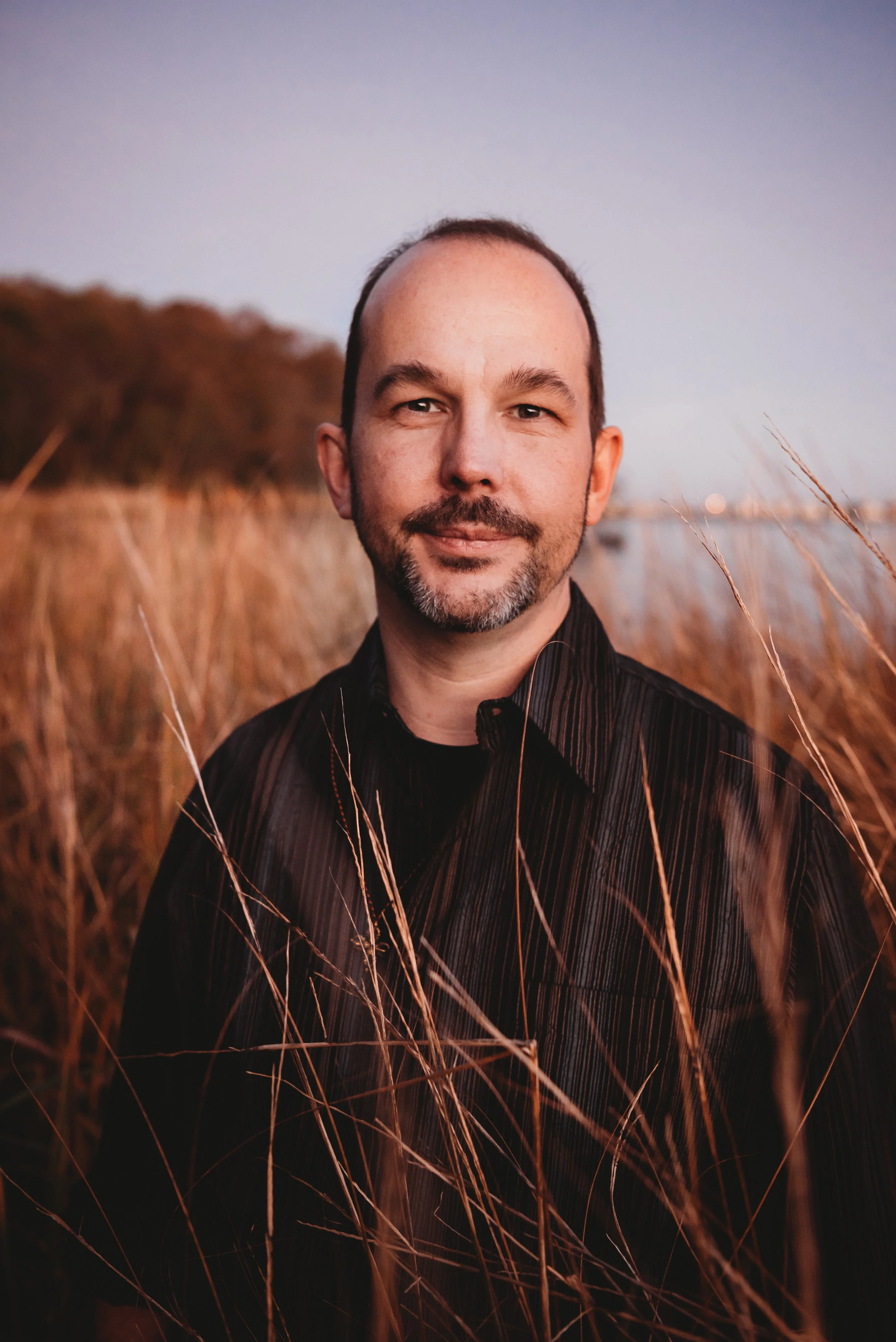 Portrait of a man with a beard and short hair, standing in tall, dry grass during sunset.
