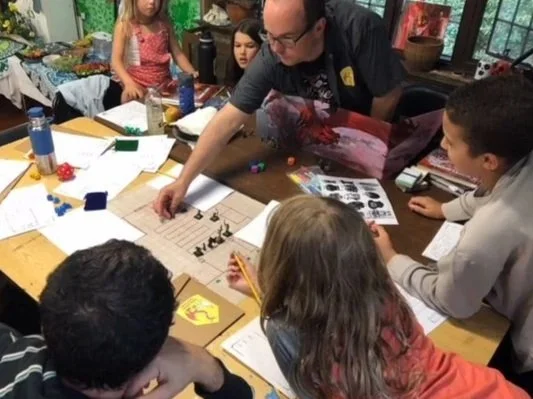 A group of children and an adult gathered around a table engaged in a tabletop game or activity, with game pieces, papers, and notebooks scattered on the table in a well-lit room with large windows.