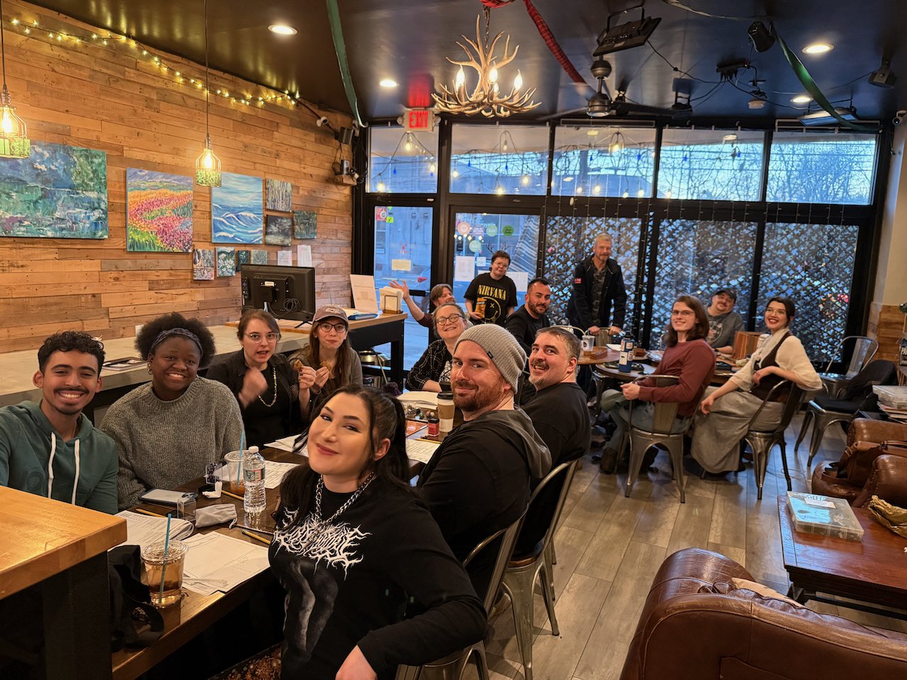 A group of smiling people sitting around tables in a cozy restaurant with wood-paneled walls, colorful paintings, and decorative lighting