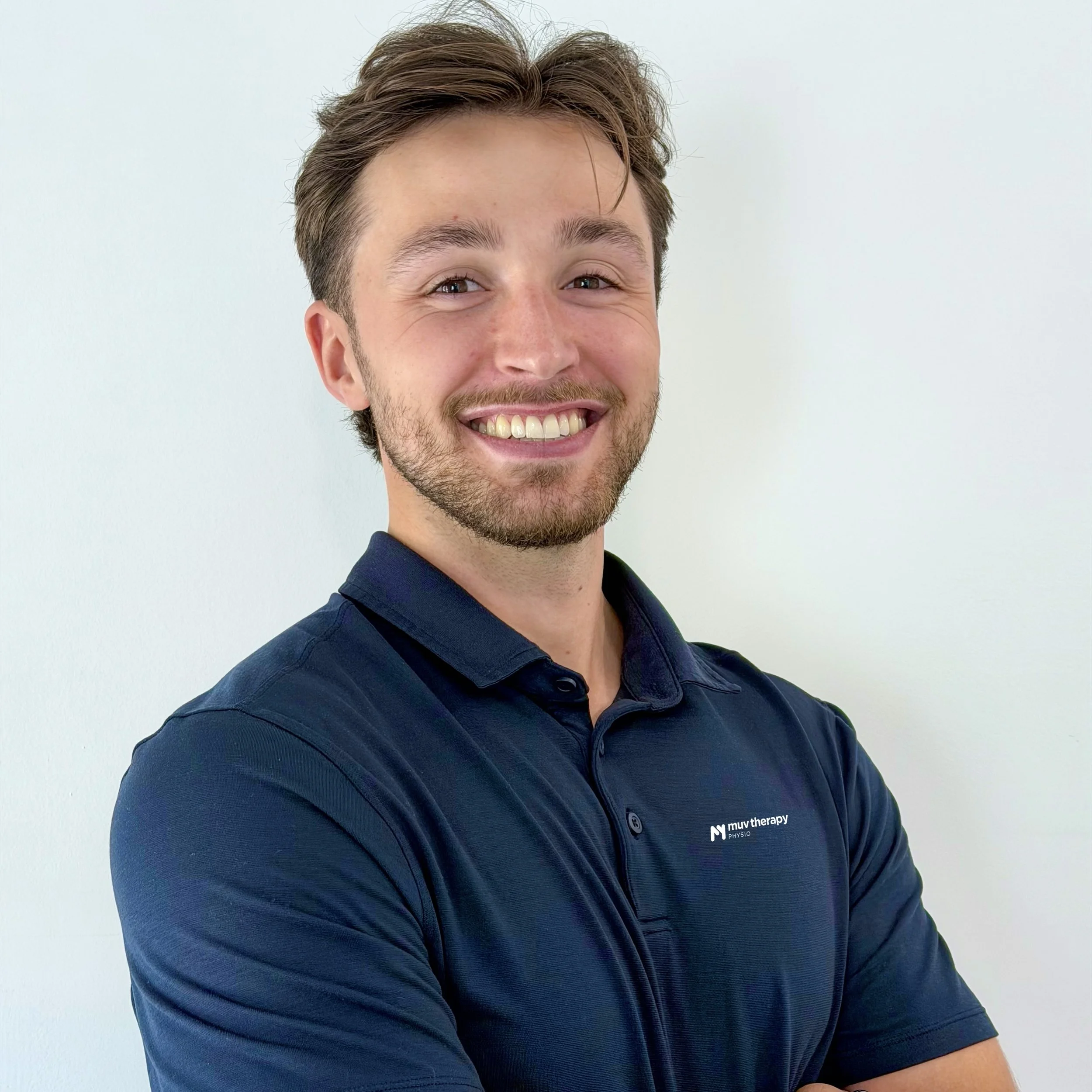 A smiling young man with brown hair and a beard, wearing a navy polo shirt, standing against a white wall.