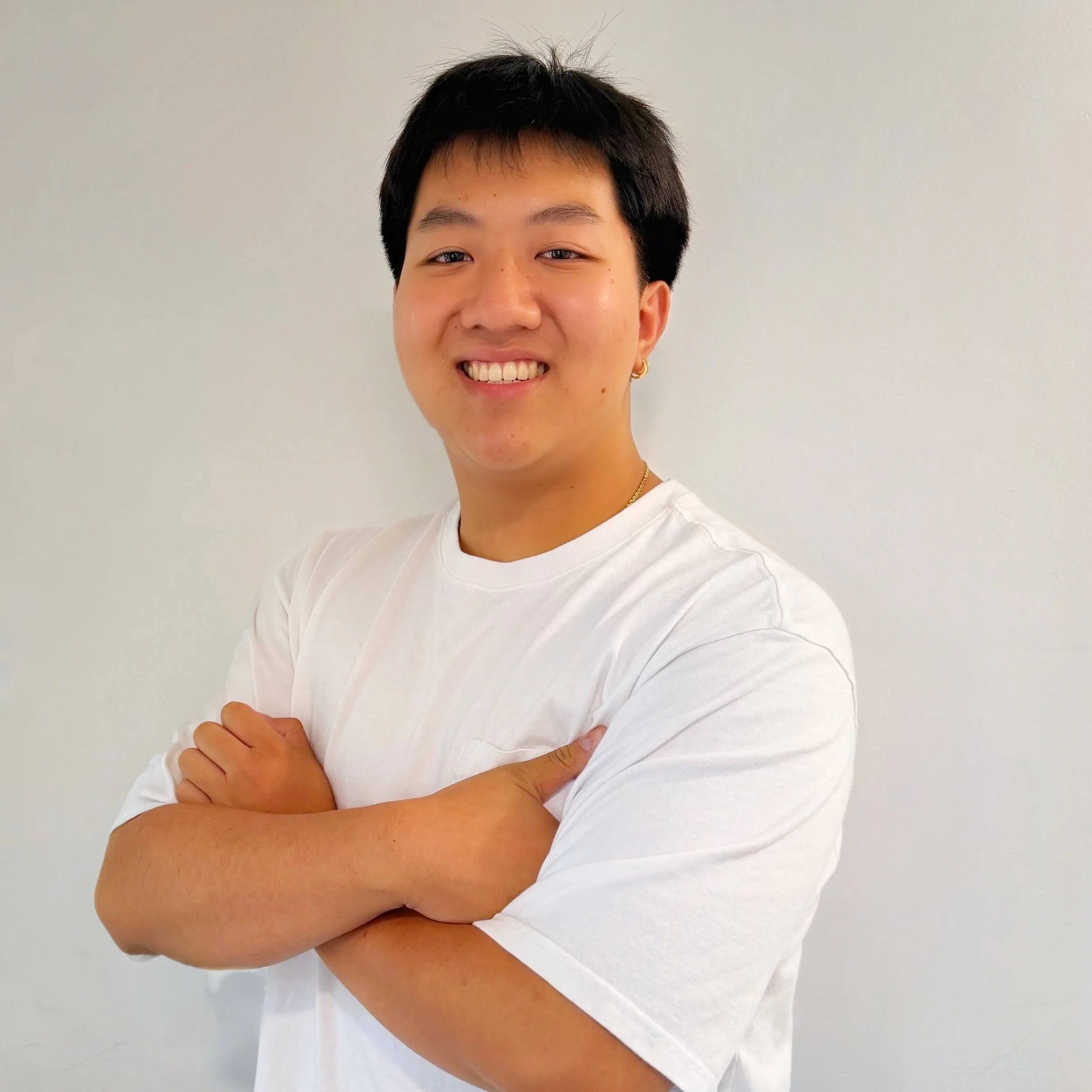 Young person with short black hair smiling, arms crossed, wearing a white t-shirt, standing against a plain light-colored wall.