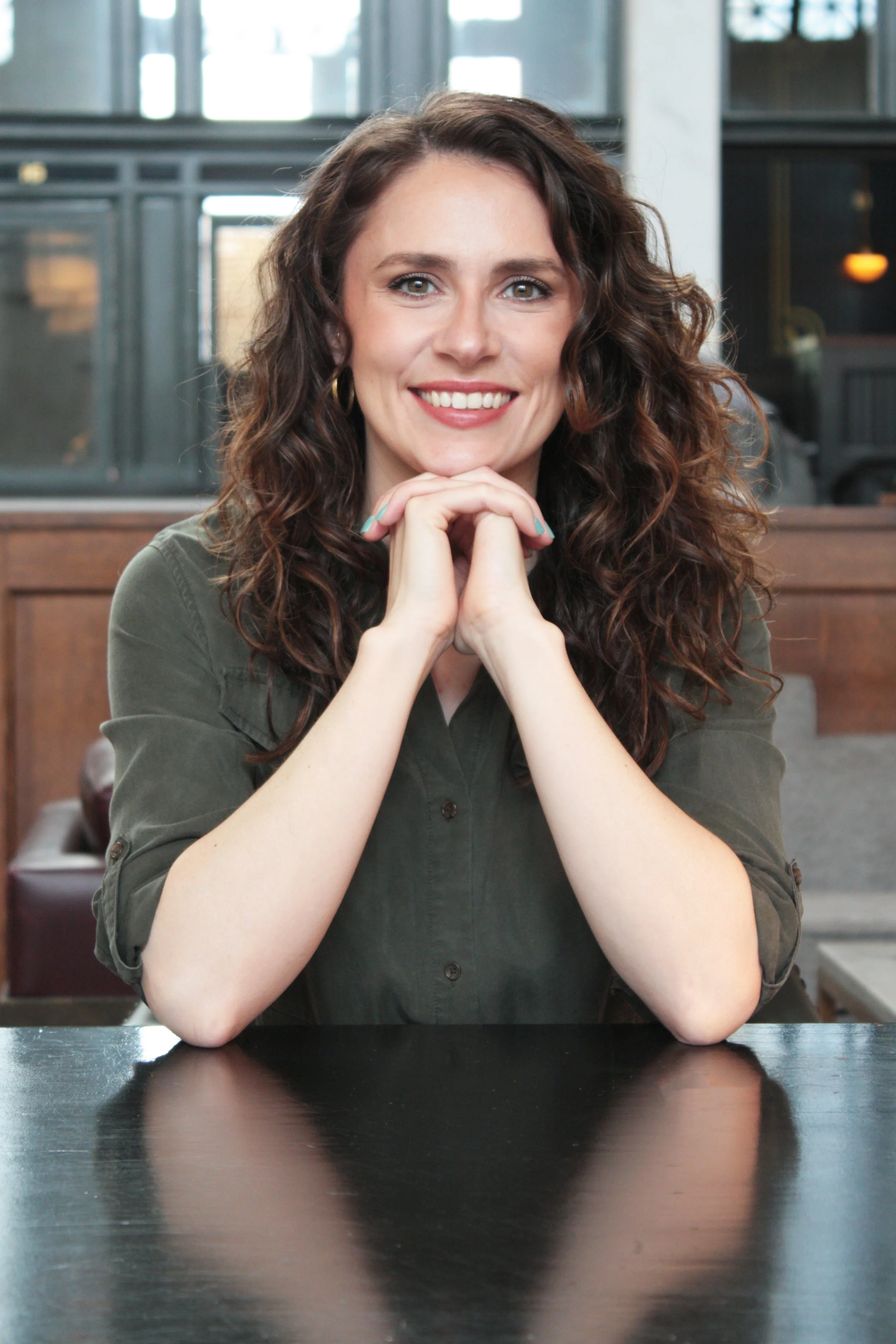 A woman with wavy brown hair and fair skin, smiling with her hands clasped under her chin, sitting at a dark table in a modern indoor setting.