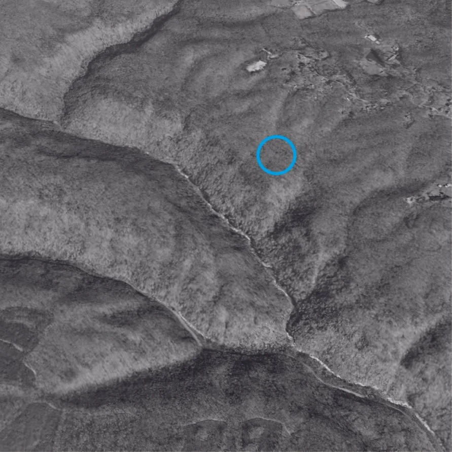 A black and white aerial view of forested hills with a small blue circle marking a location.