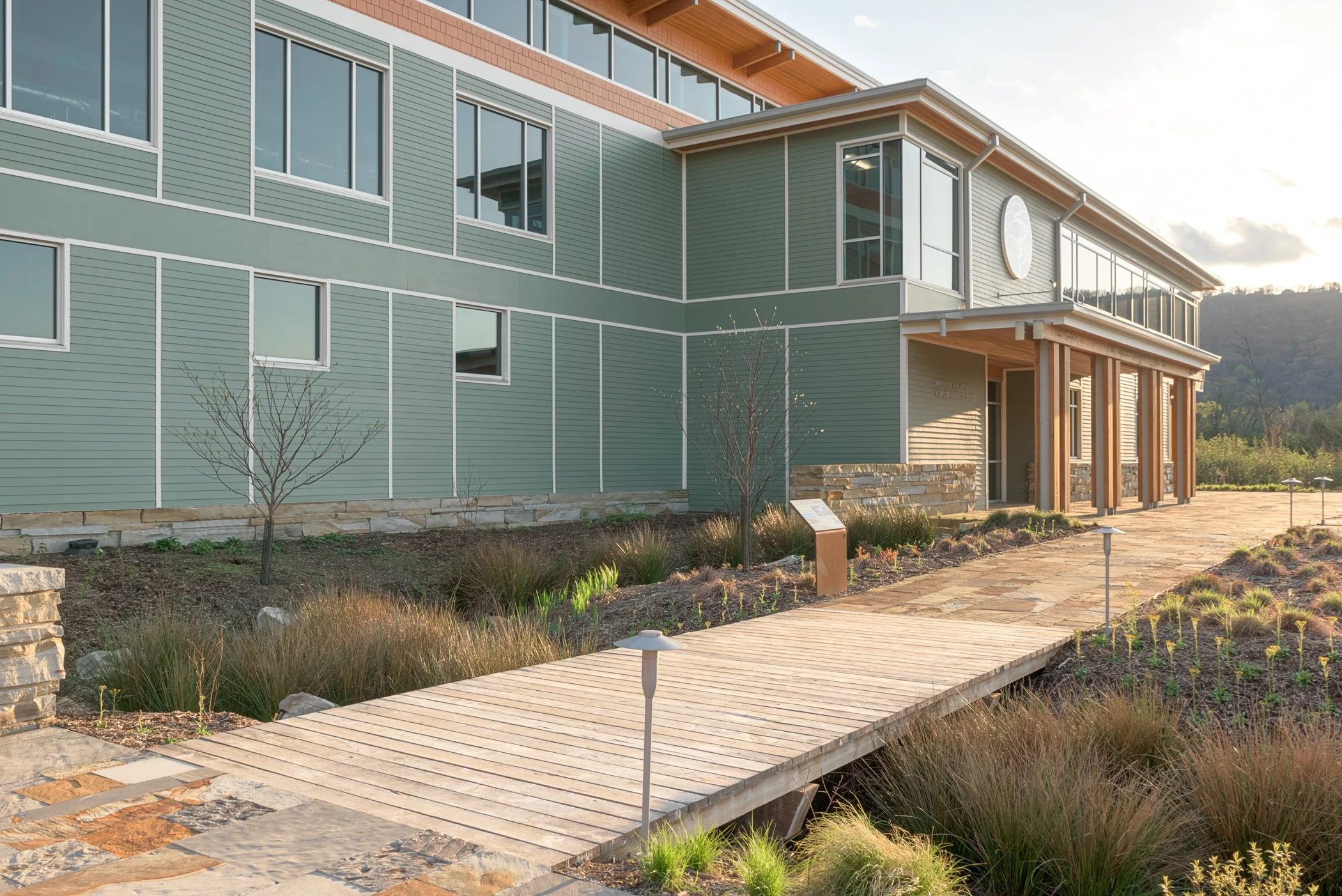 Exterior of a modern building with green siding, multiple windows, and a stone and wood entrance. A wooden walkway with small pathway lights leads to the entrance, surrounded by landscaped plants and small trees.