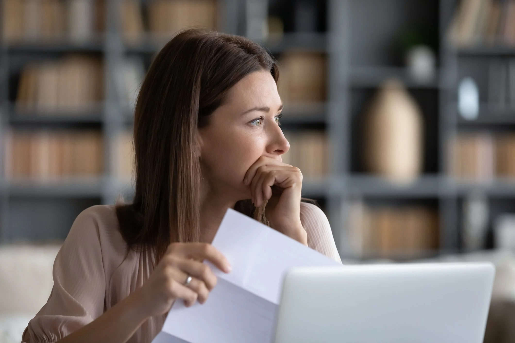 A woman sitting at a desk holding a paper, looking thoughtfully to the side with a laptop open in front of her, in a room with a bookshelf behind her.