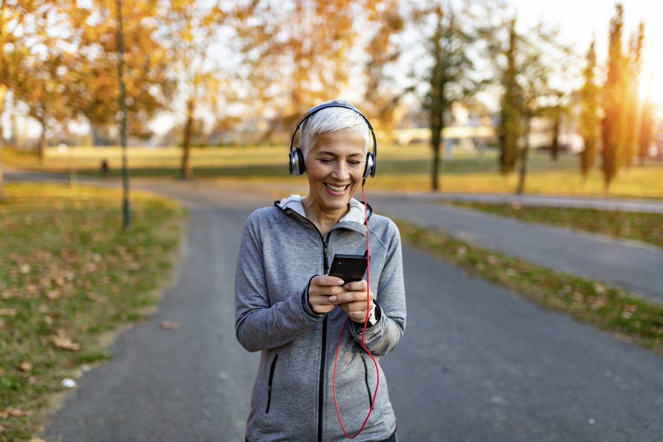 An elderly woman with short gray hair wearing a gray athletic jacket and headphones, smiling while looking at her phone in a park during autumn sunset.