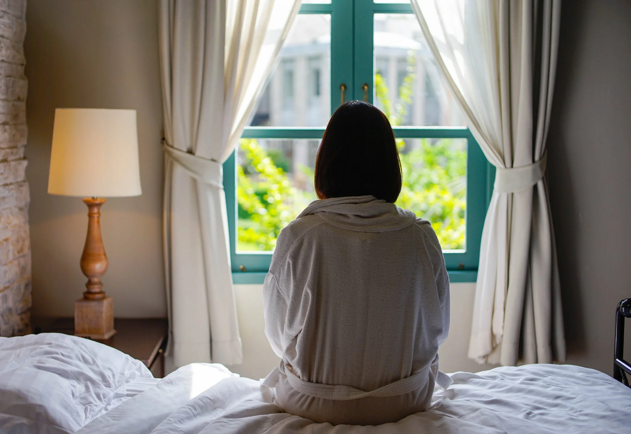 A woman sitting back to on a bed looking out a window in a dark bedroom