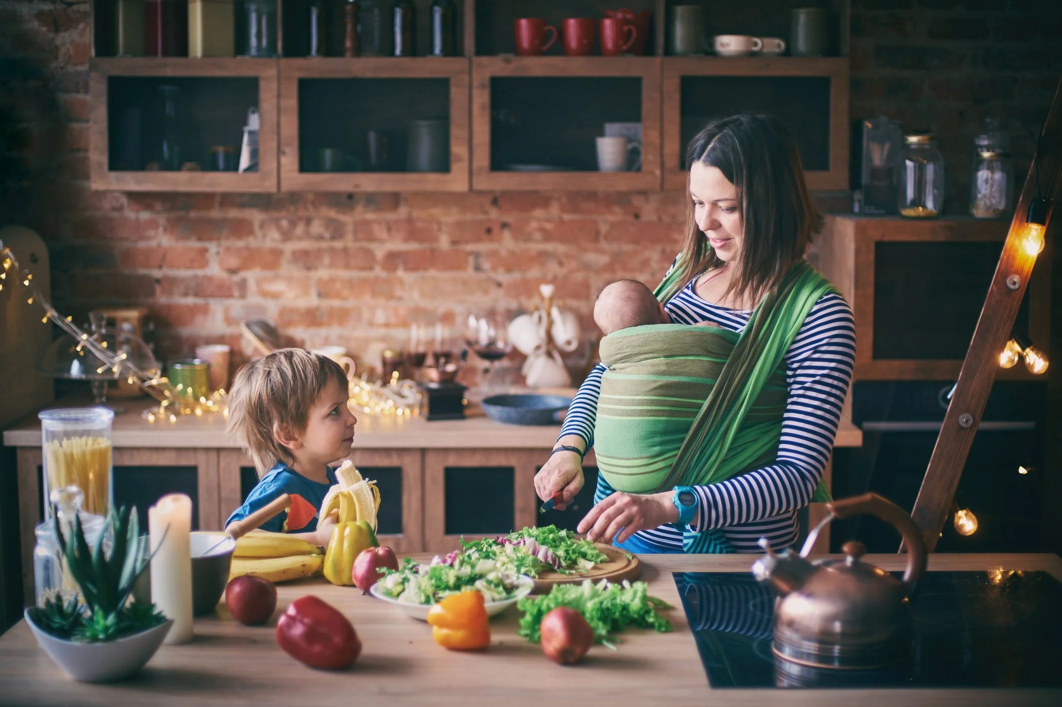 A woman with a toddler and holding a baby while in a kitchen preparing food