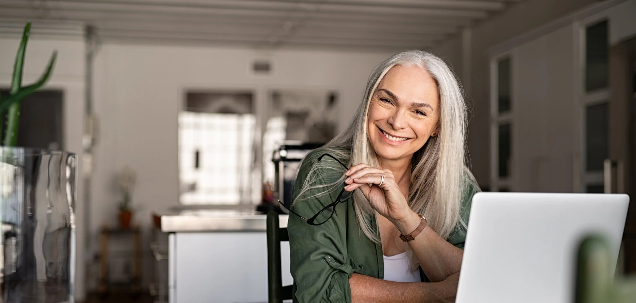 Mature white haired woman smiling while sitting at a table with her laptop open