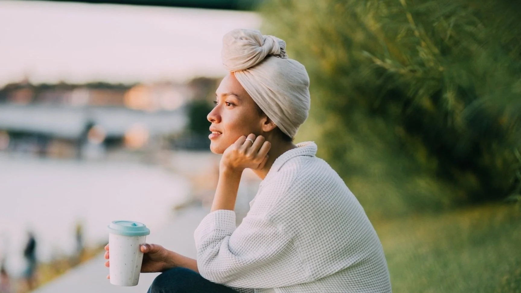 Woman sitting outdoors near water, holding a reusable coffee cup, wearing a light-colored head wrap and a white sweater, gazing peacefully into the distance.