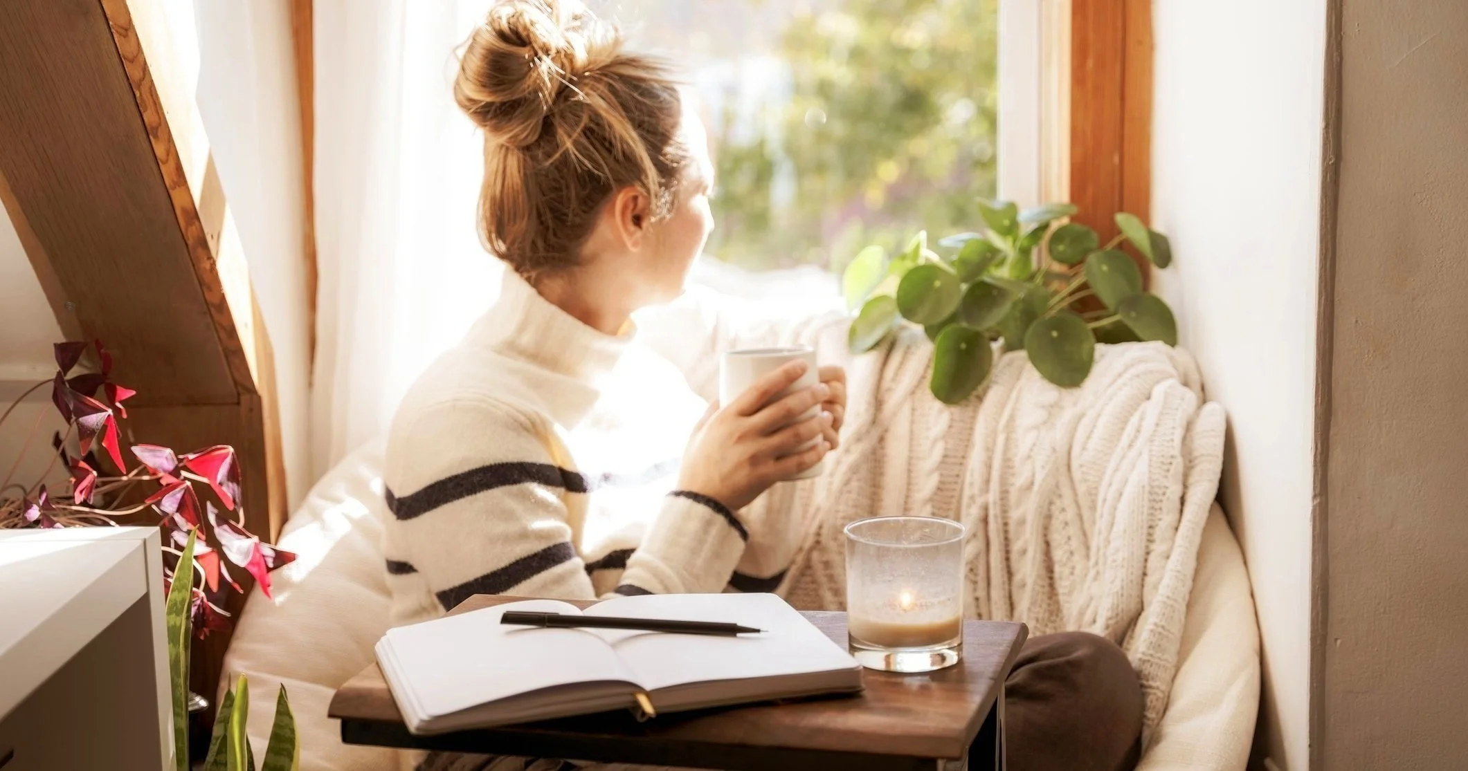 Woman sitting outdoors near water, holding a reusable coffee cup, wearing a light-colored head wrap and a white sweater, gazing peacefully into the distance.