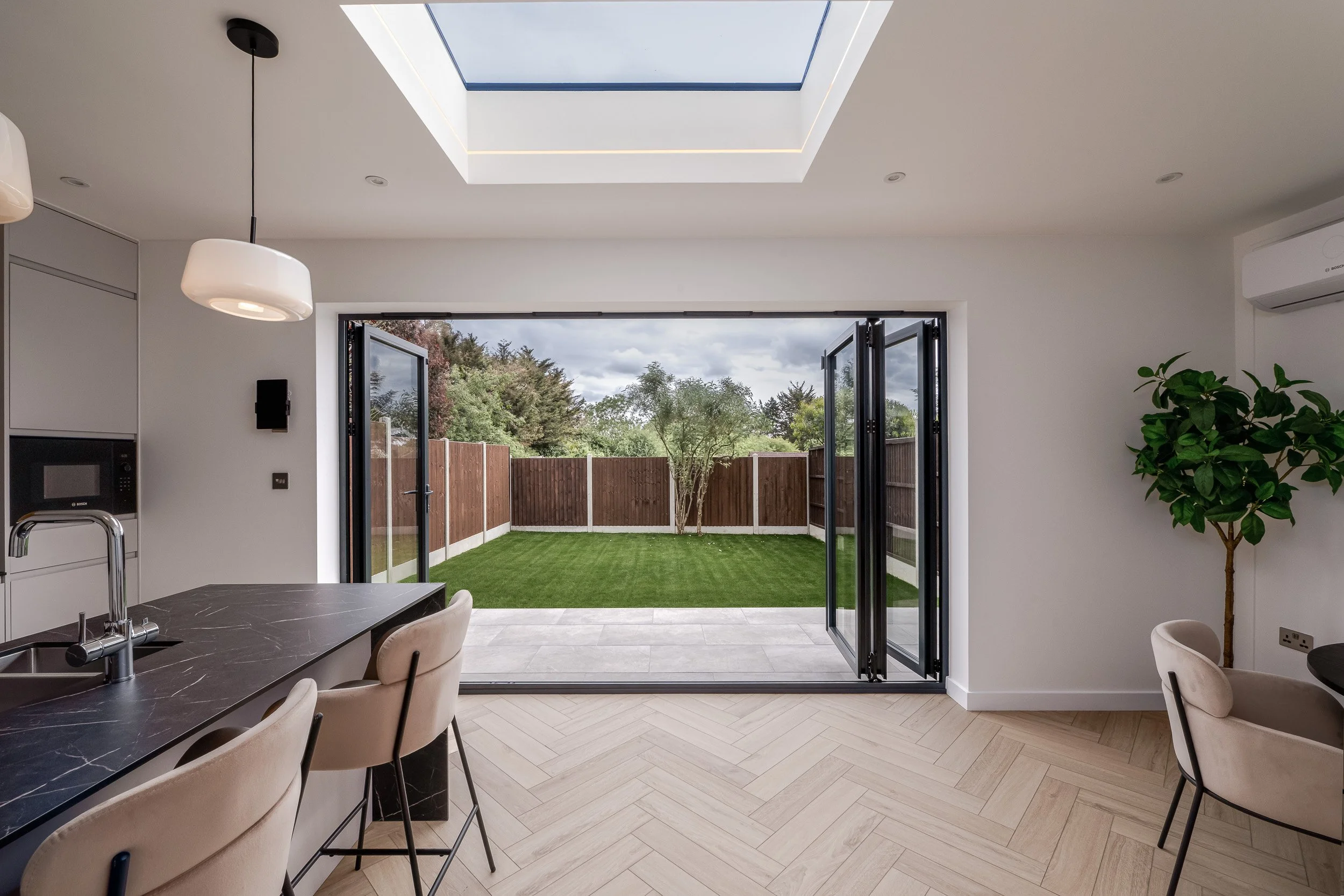 Architectural photograph of a modern residential kitchen extension, showcasing open-plan indoor-outdoor living. The wide black aluminium bifold doors are pulled completely open, creating a seamless transition from the indoor herringbone wood flooring