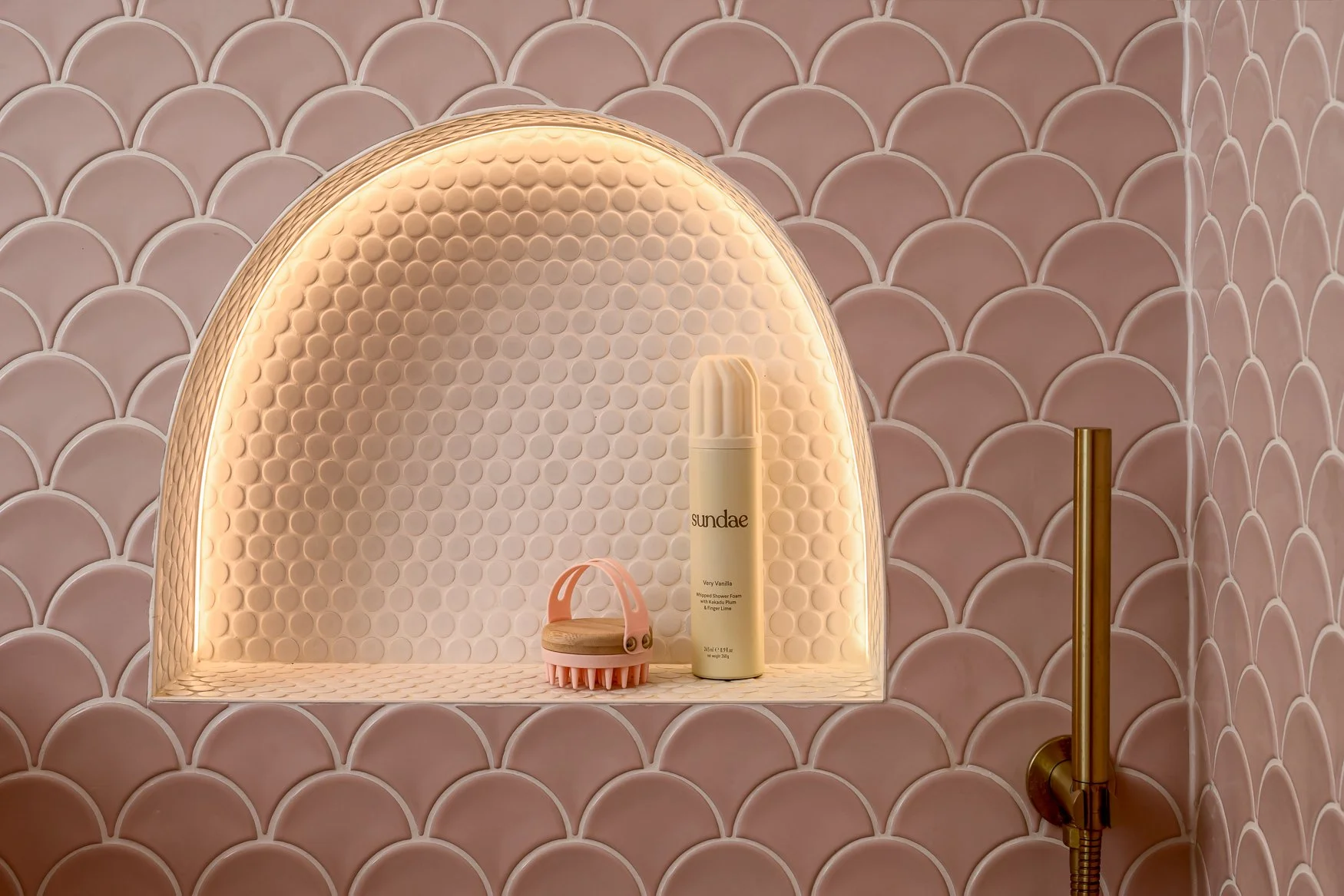Pink scalloped shower tiles with brass shower fittings and built-in illuminated niche.
