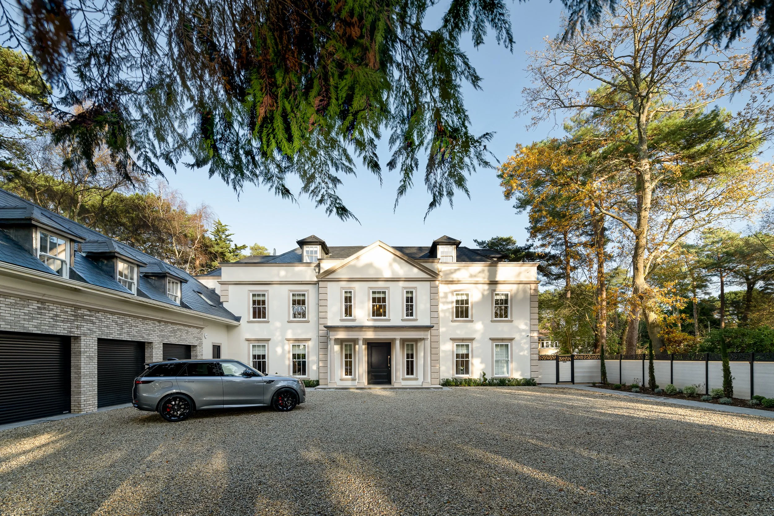 Luxury residential architecture photography in Poole, showing a classical-style home with a gravel driveway, garage building and landscaped trees in soft natural daylight.