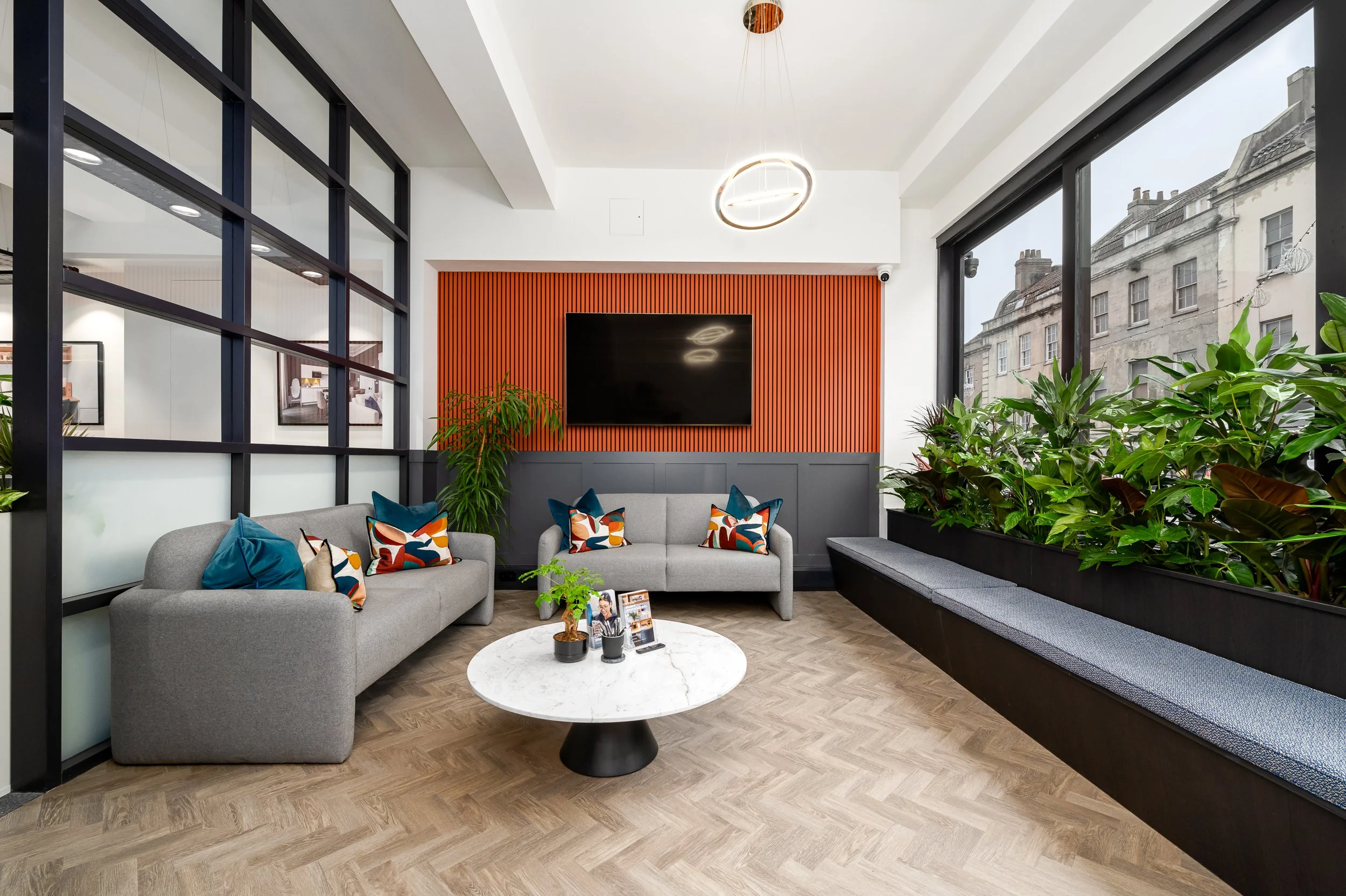 Modern lounge area with gray sofas, colorful throw pillows, a round white marble coffee table, tall green plants, large windows, a flat-screen TV mounted on a wooden slat accent wall, and contemporary lighting fixtures.