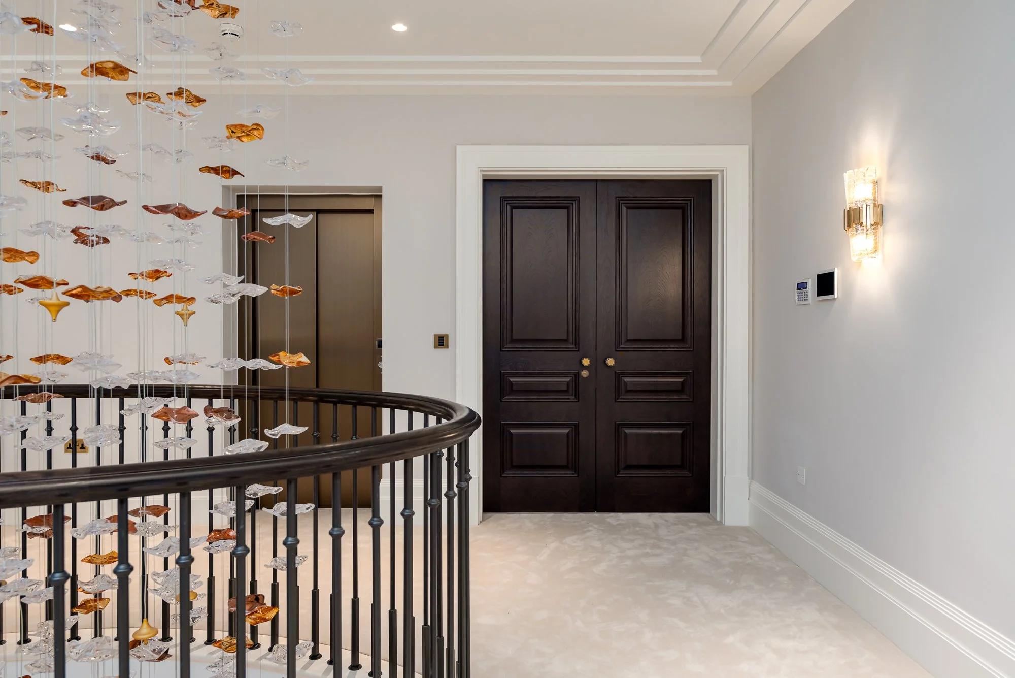 A luxury landing with dark timber double doors set within white architectural trim. The image highlights the rich door finish, classic panel detailing, curved balustrade and decorative glass chandelier, showing the doors as part of a high-end residen