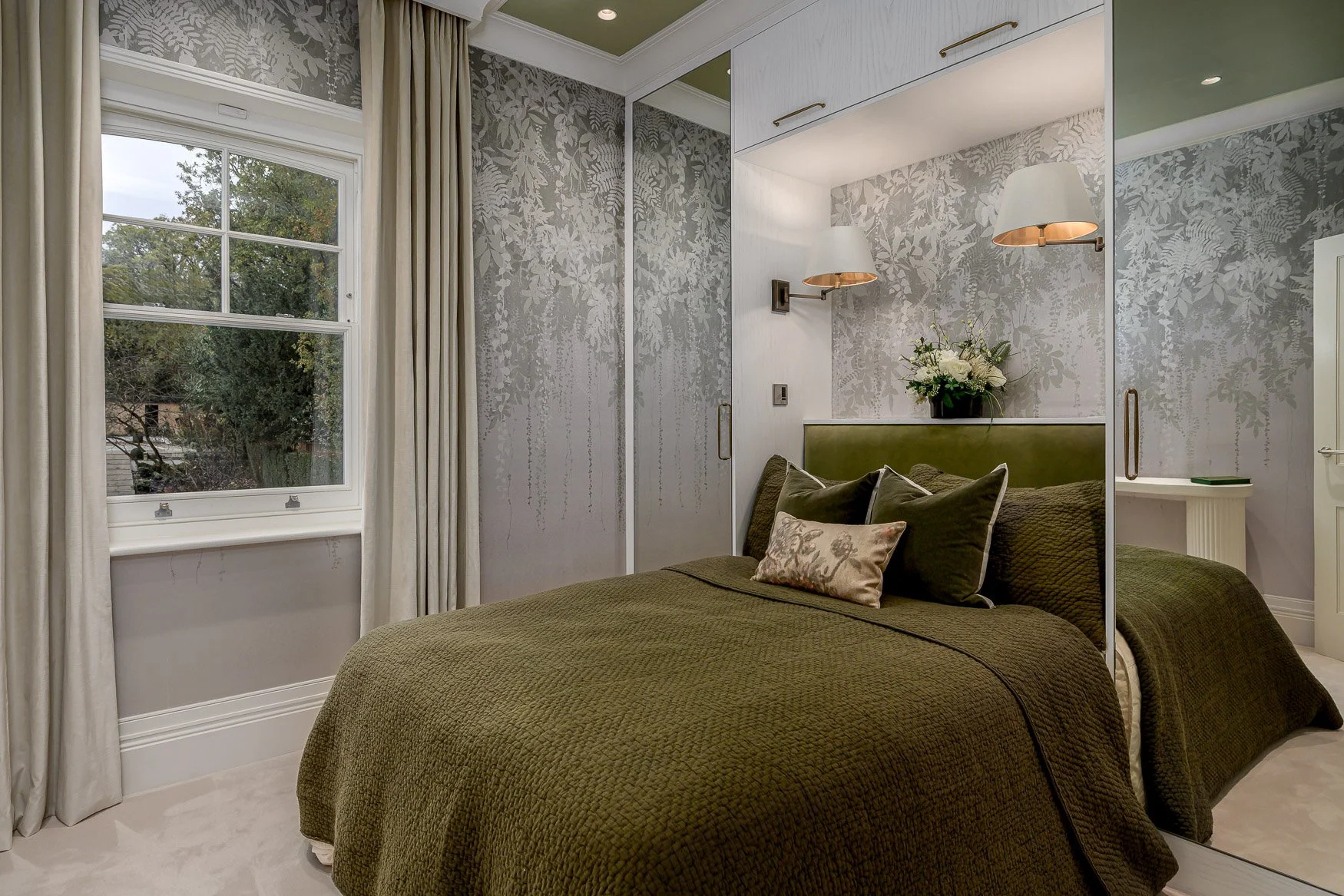 A cozy bedroom with a large window, cream curtains, and a bed with dark green bedding, pillows, and a headboard. There is a floral arrangement on the wall behind the bed and two modern wall-mounted lights.