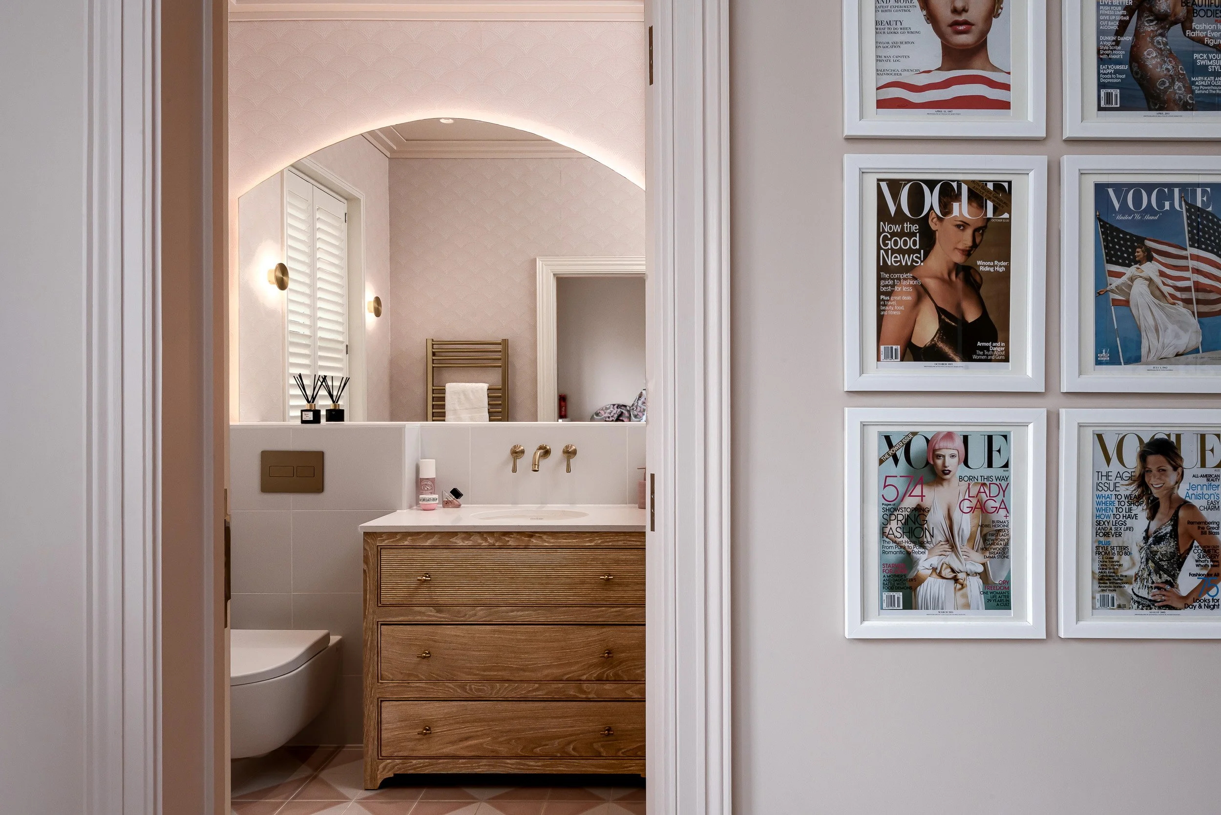 A soft pink bathroom photographed in London, featuring an arched mirror, warm wall lighting, brass taps, a wooden vanity unit, pale tiles and framed fashion magazine covers in the adjoining hallway. The image captures a feminine, polished bathroom de
