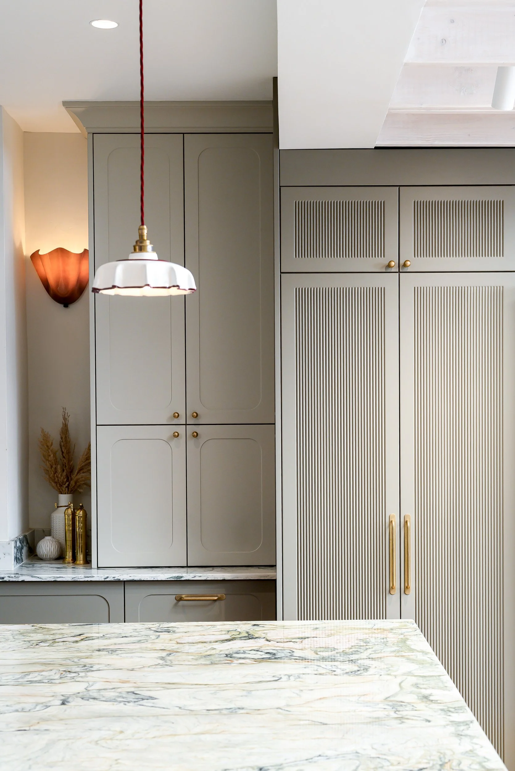 Interior of a kitchen featuring beige cabinetry with gold hardware, a marble countertop, decorative vases, a wall-mounted sconce, and a hanging pendant light.