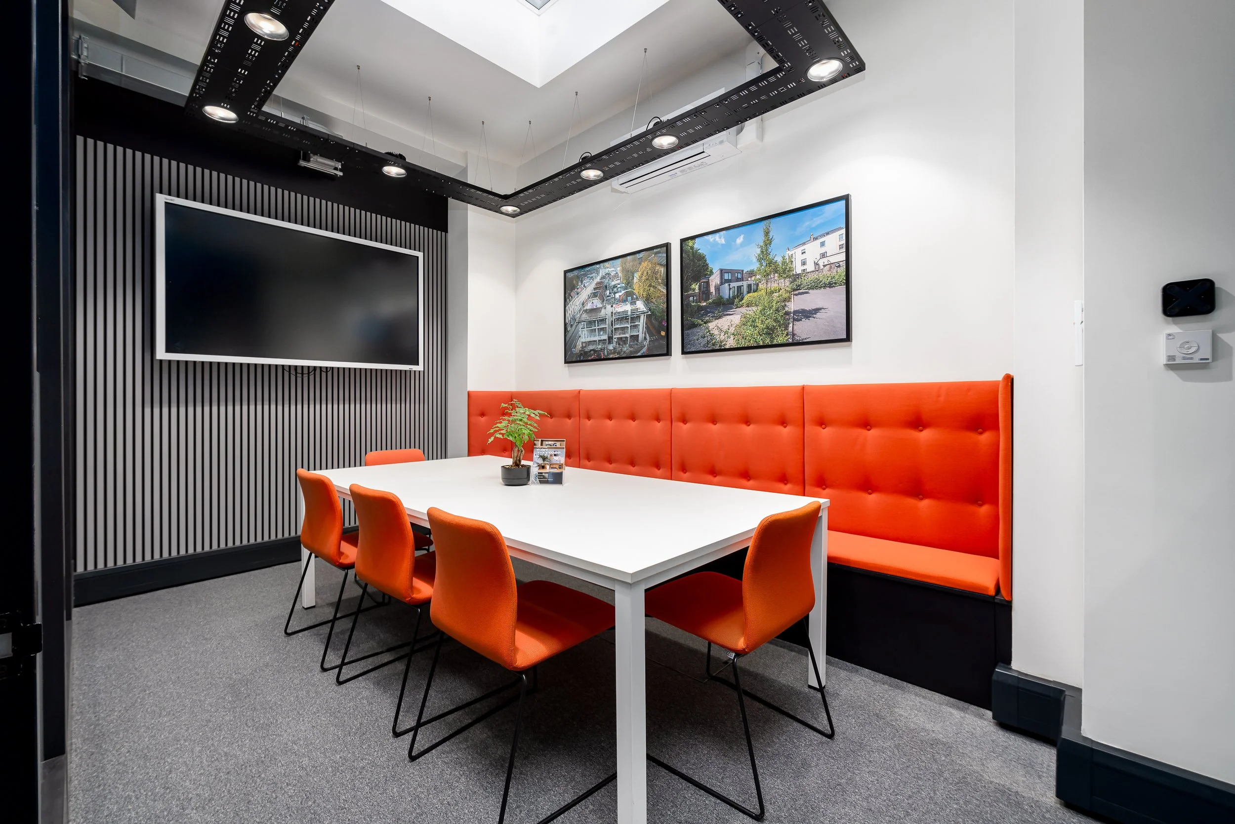 A modern meeting room with a white table, orange chairs, a red upholstered bench, wall-mounted screens showing outdoor scenes, and a decorative black ceiling fixture with integrated lights.