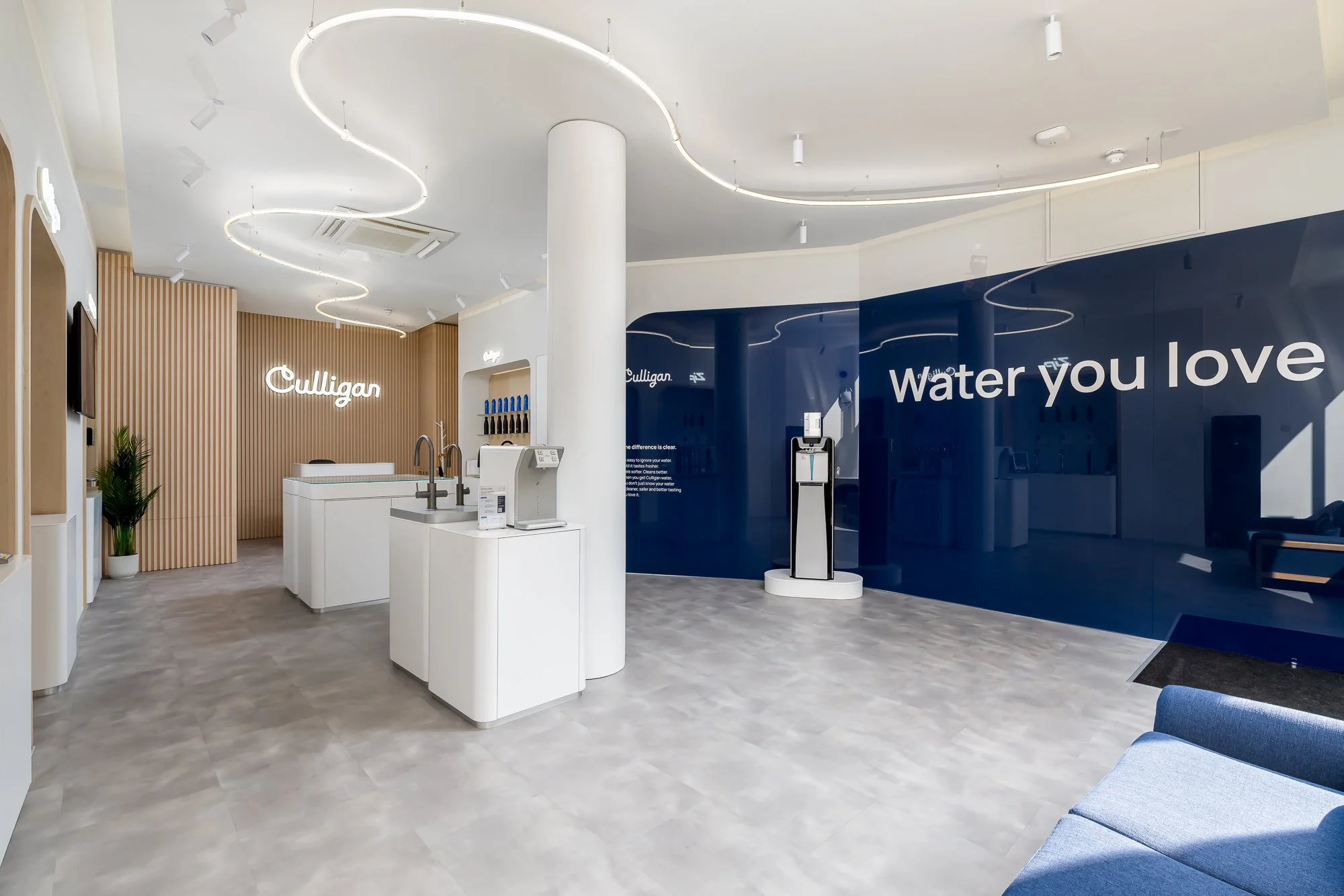 Modern interior space with white and wood accents, featuring a sign that reads 'Culligan', a water station, and a wall with the words 'Water you love' in white text on a dark blue background. The ceiling has stylish curved light fixtures and several 