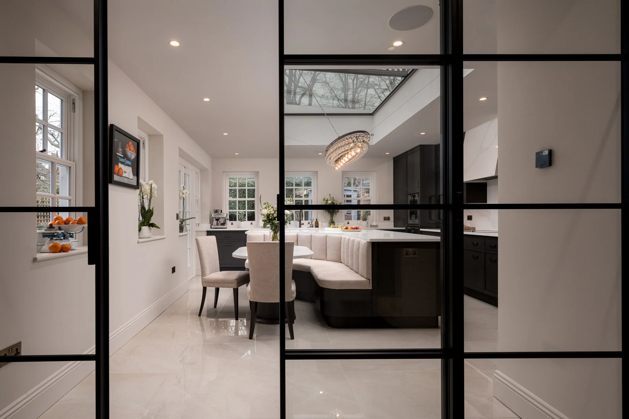 Interior architecture photograph looking through black-framed internal glazing into a residential kitchen with rooflight, island seating and polished stone flooring.