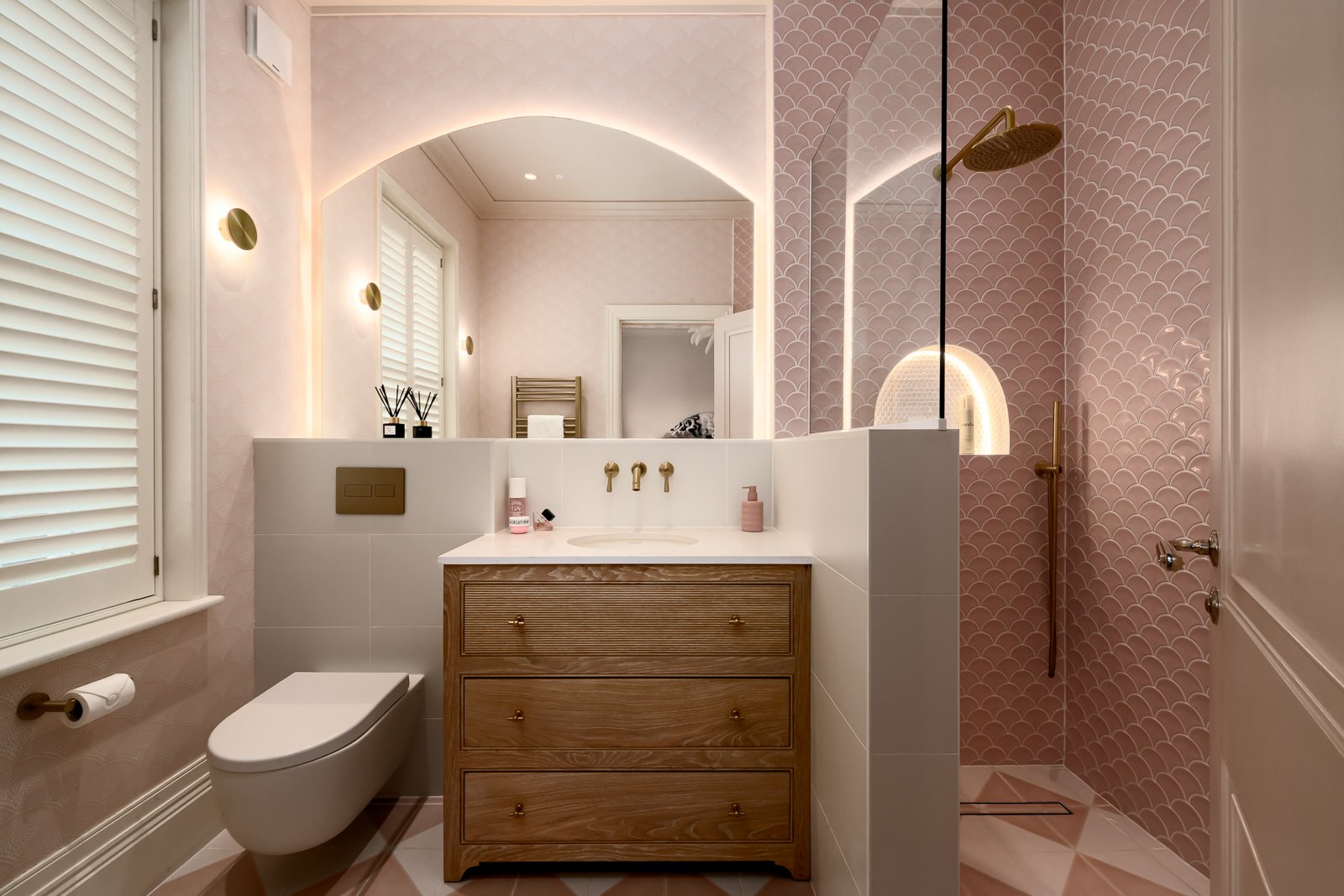 A modern bathroom with a pink and white color scheme, featuring a walk-in shower with pink scalloped tiles, a wooden vanity with drawers, a white toilet, a large mirror with backlighting, and window shutters. London i nterior design project