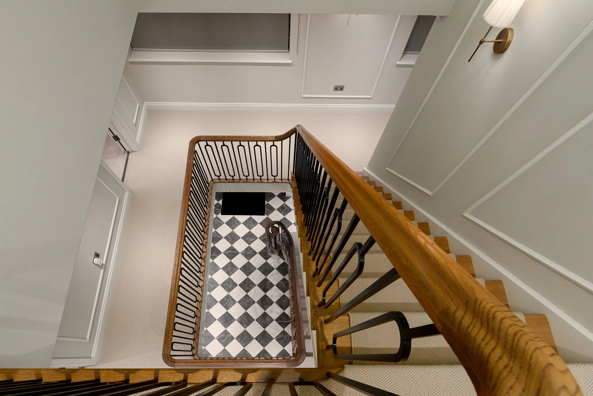 Interior architecture photograph looking down a residential staircase with timber handrail, black metal balustrade and checkerboard stone hallway floor.