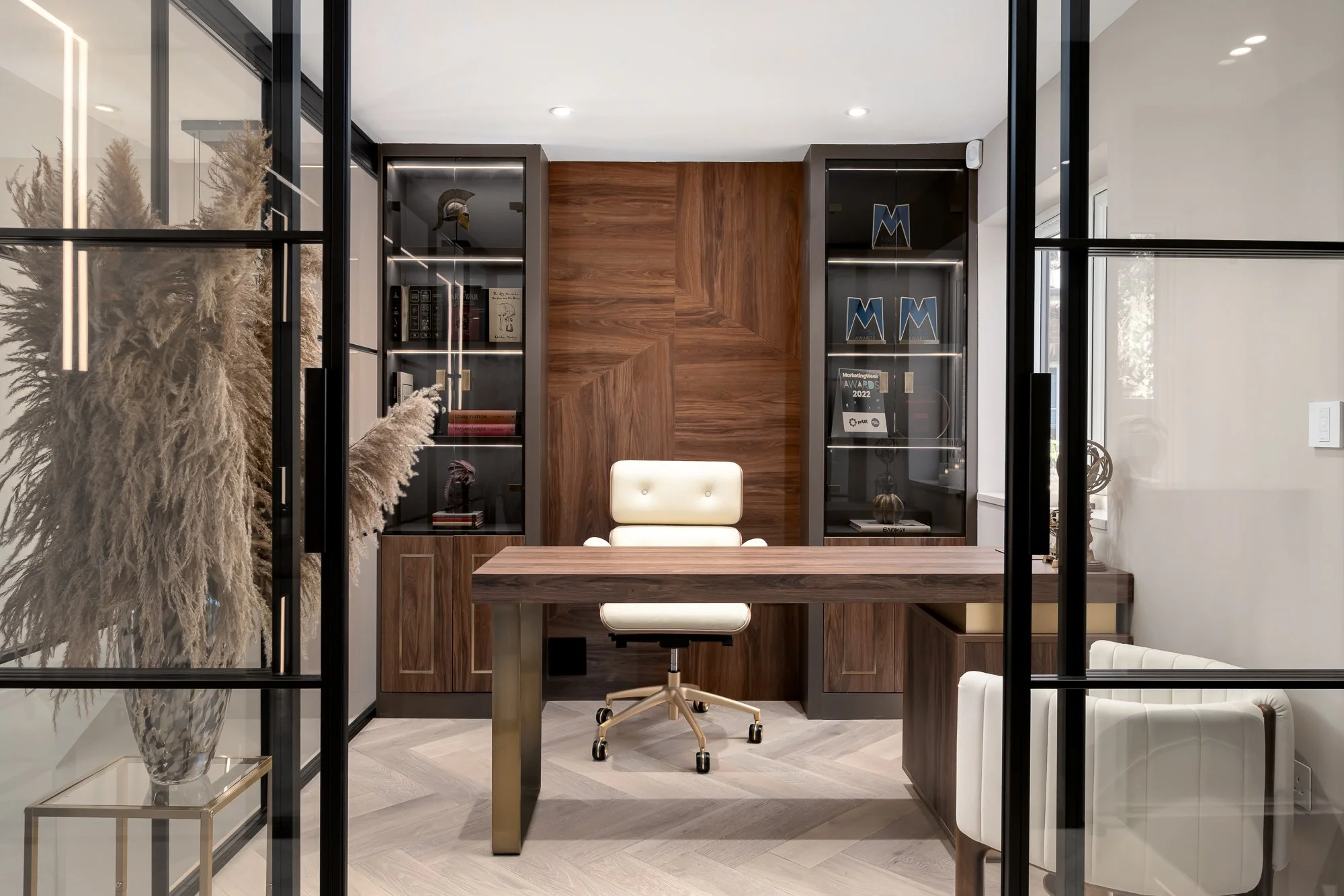 A close view through black framed glass doors into a luxury home office with bespoke timber joinery, built-in shelving and soft neutral finishes. The image highlights the slim metal frame, glazed door system and how the doors create separation withou