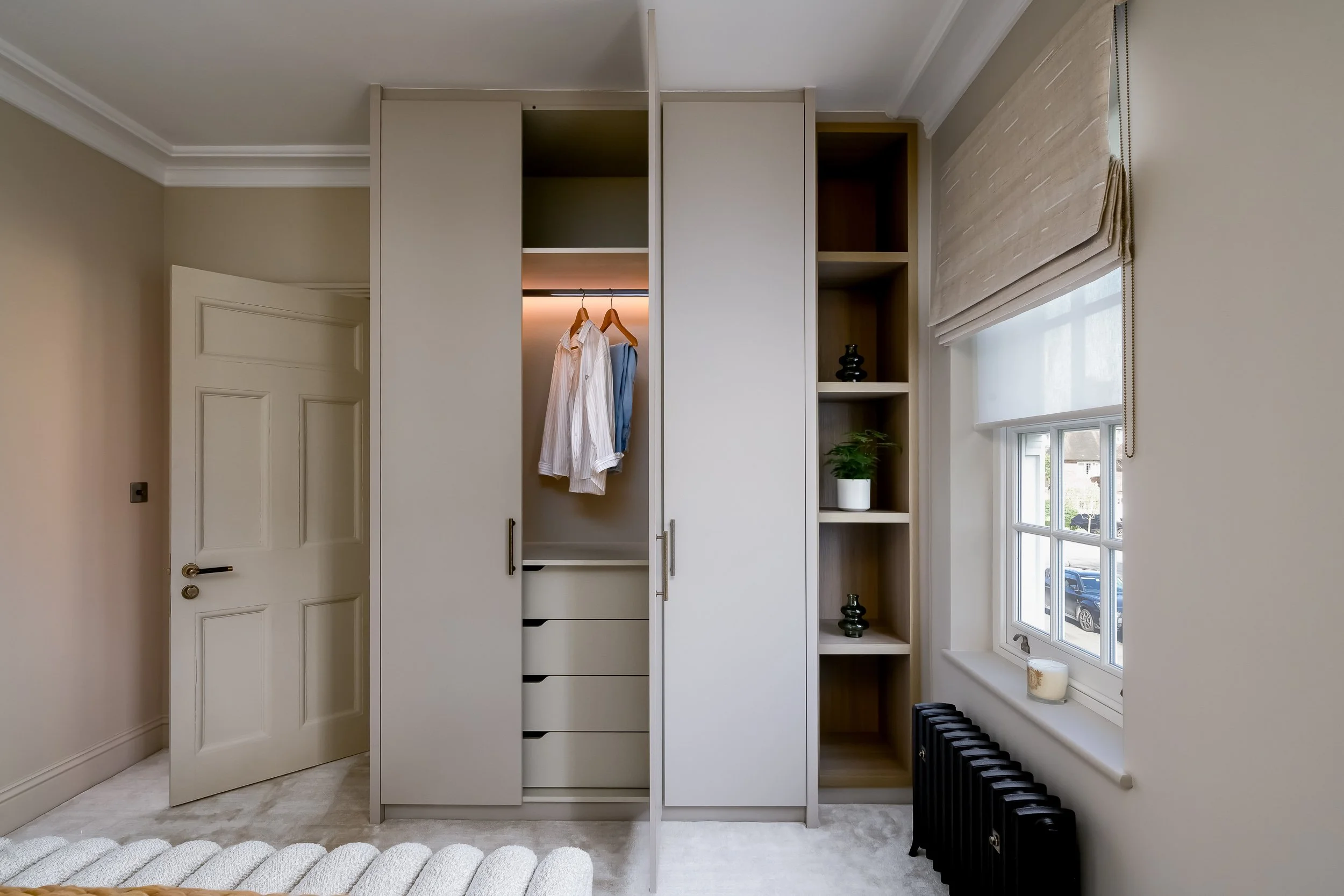 A closet with beige walls, a white door, and built-in storage including drawers and open shelves. Two shirts hang inside the closet, with a window on the right side of the room allowing natural light. There is a black radiator below the window, and a candle on the windowsill.