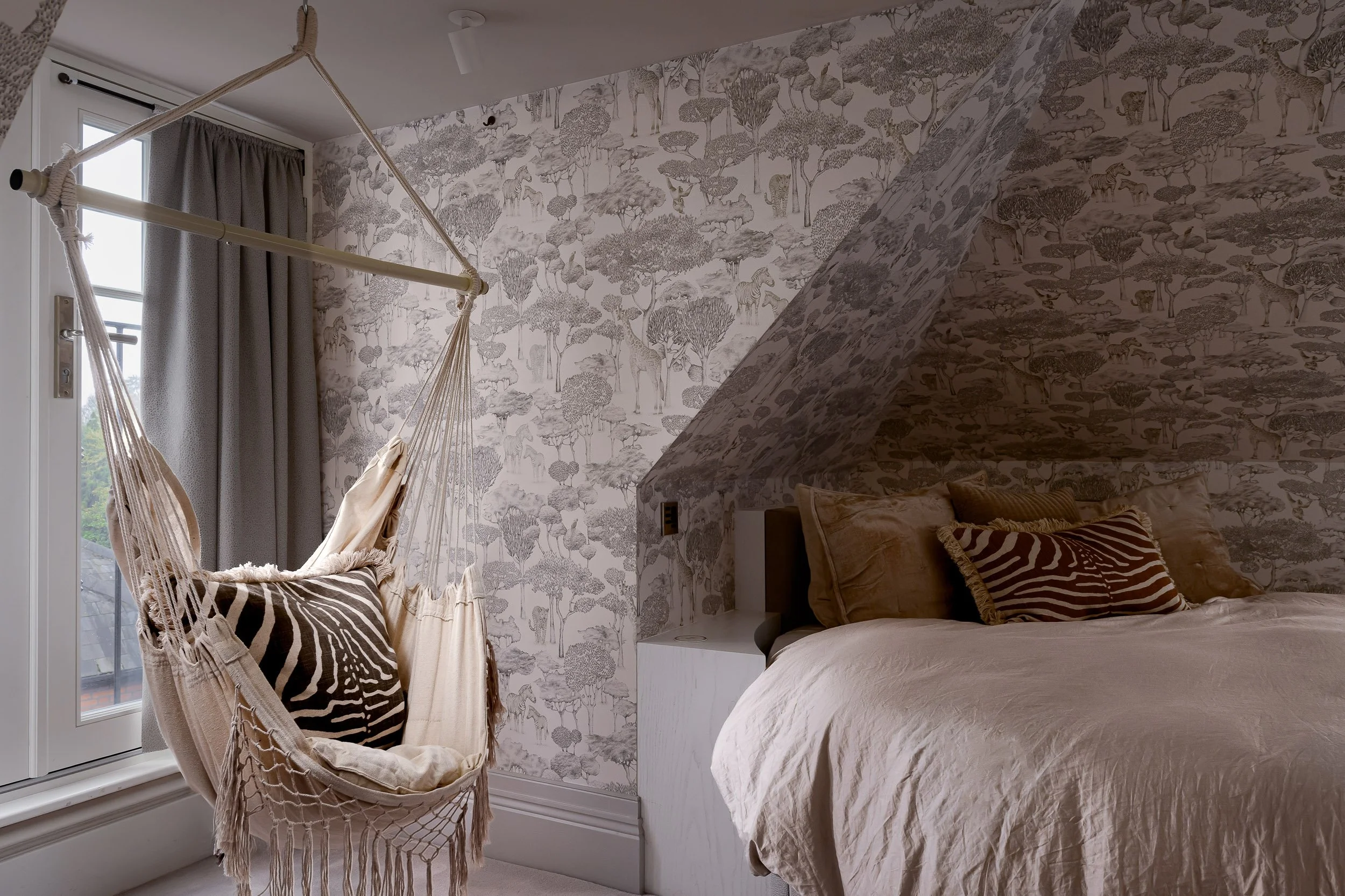 A teenager’s bedroom photographed in London, featuring patterned wallpaper, a suspended hammock chair, soft neutral bedding, layered cushions, natural textures and gentle window light. The image captures a relaxed, personal space with a mix of playfu