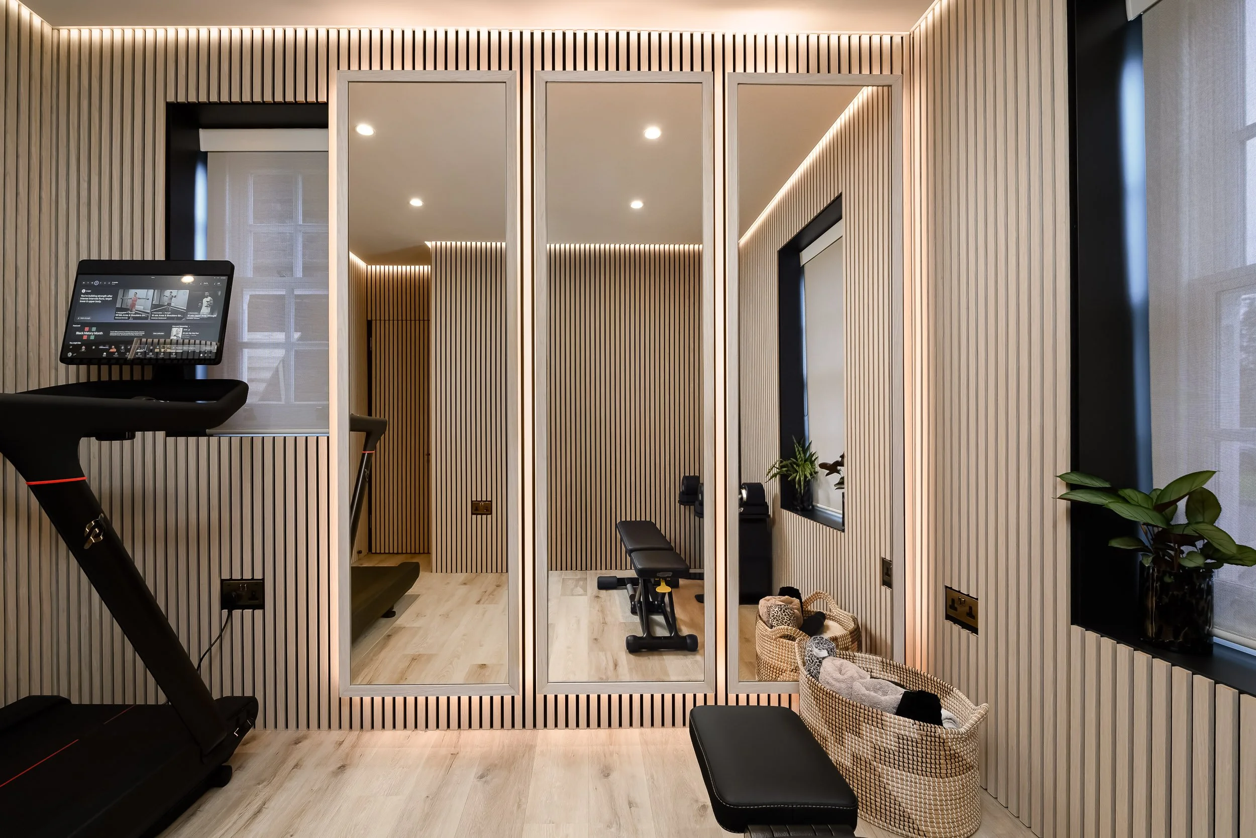 A modern wellness or fitness room with wood slat walls, a treadmill on the left, and a small seating area with benches and baskets of clothes or towels. The room is well-lit with recessed ceiling lights and LED strip lighting along the ceiling edges.