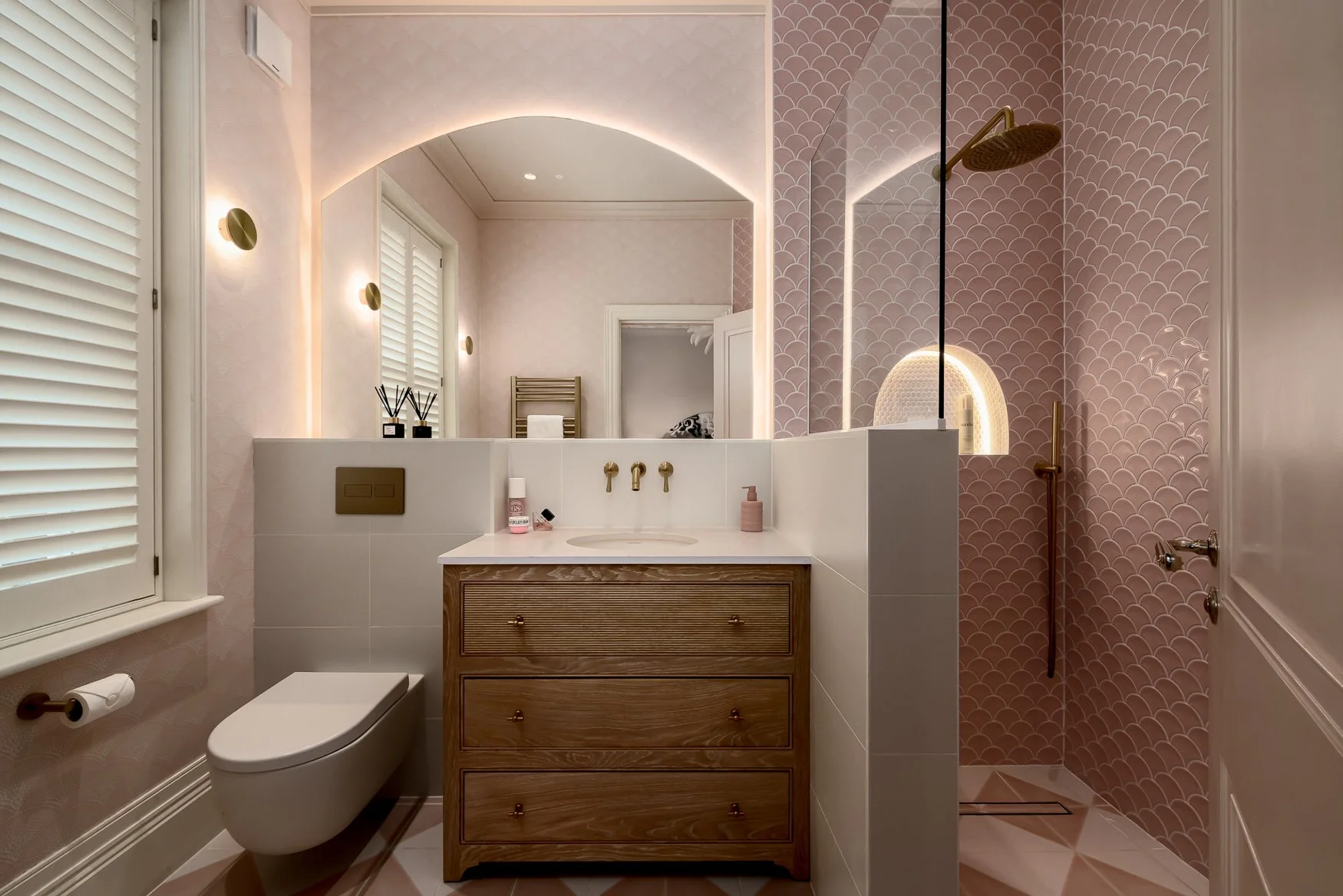Ensuite bathroom vanity with arched mirror, brass wall taps and pink tile details.